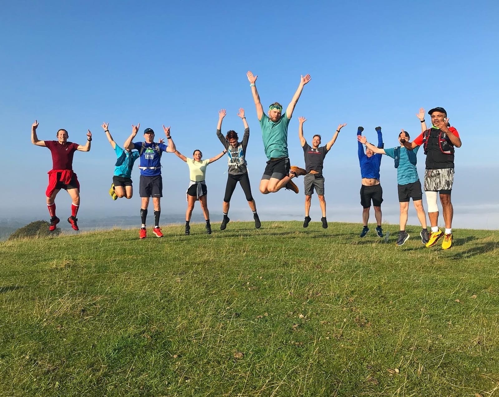A group of ten people jumping in the air with their arms raised on a grassy hill under a clear blue sky. They are wearing various colorful athletic outfits and seem to be enjoying an outdoor activity together.