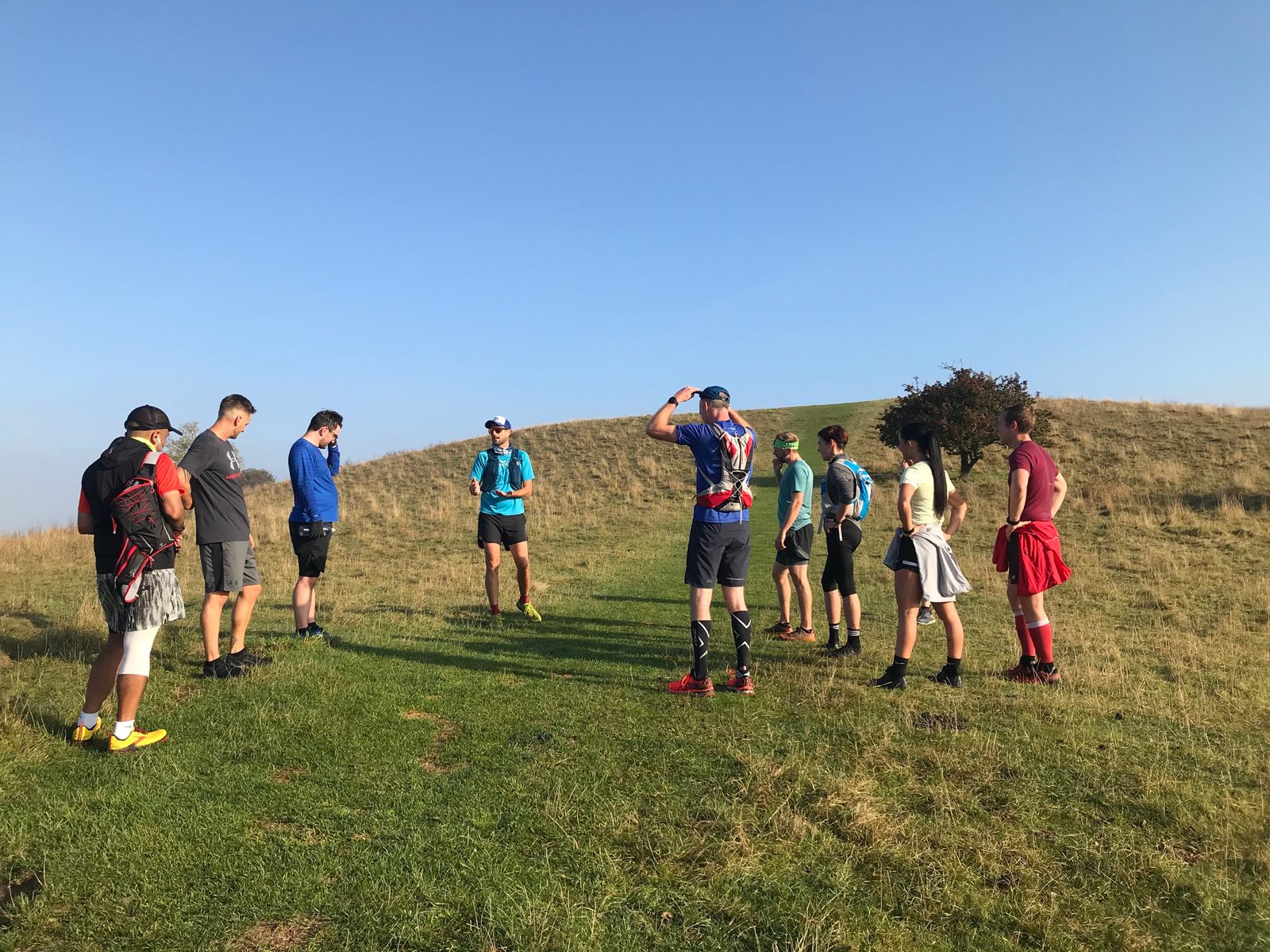 A group of nine people in athletic gear are standing on a grassy hill under a clear blue sky. Some are looking at a person in a blue shirt in the center, who appears to be giving instructions. The land is open, with short grass and sparse vegetation.