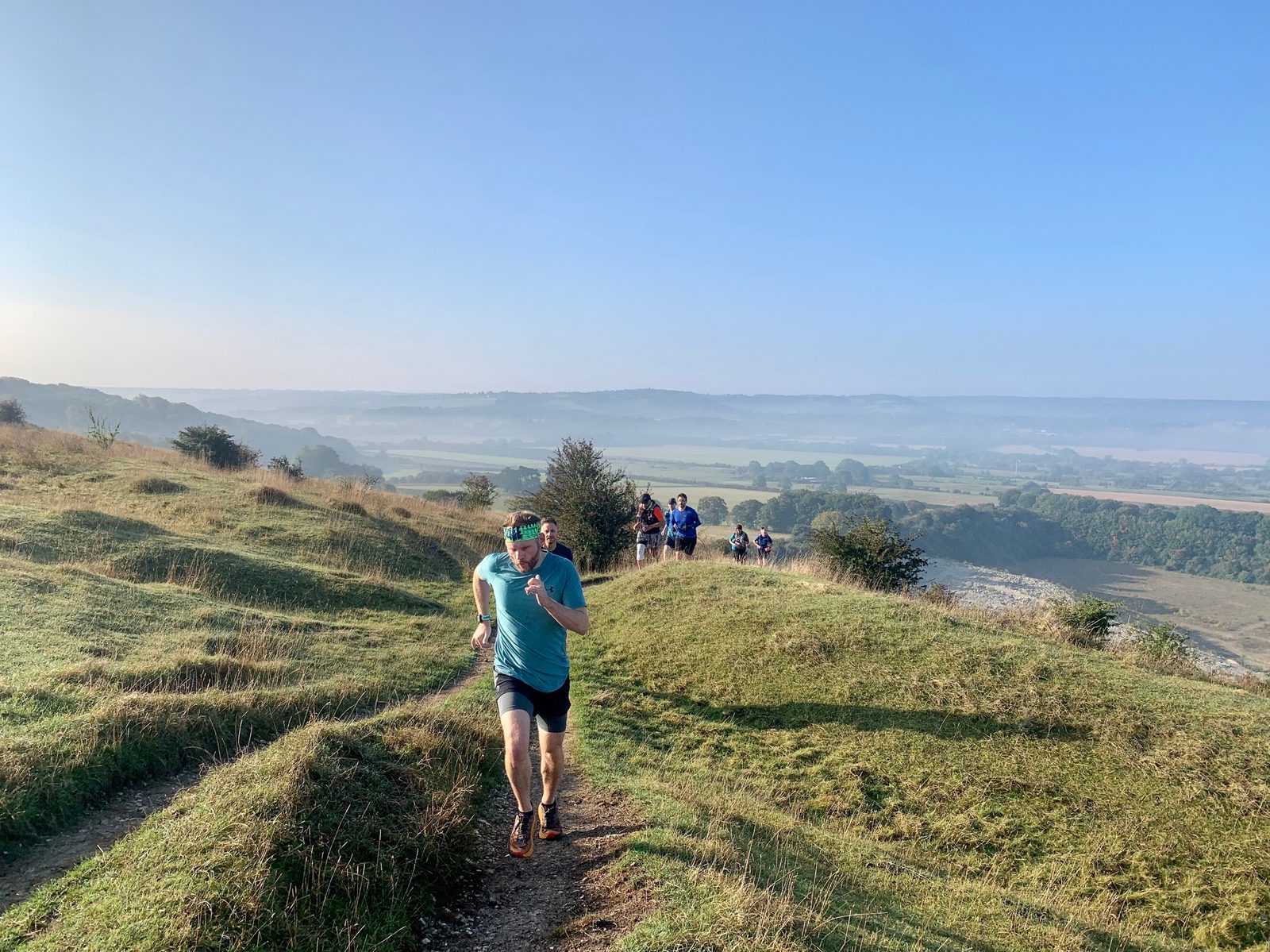 A group of runners is seen jogging up a grassy hill under a clear blue sky. The foreground shows a man in a teal shirt and black shorts running, with several other runners in the background. The landscape below stretches out with fields, trees, and slight morning mist.
