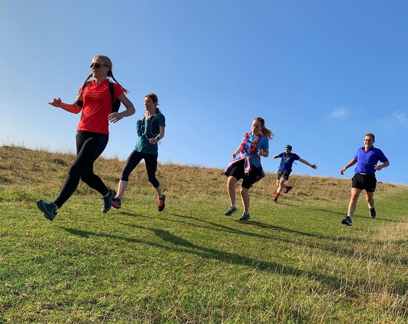 A group of five people, three women and two men, are running down a grassy hill under a clear blue sky. They appear to be participating in an outdoor activity, wearing athletic clothing and looking focused and energized.