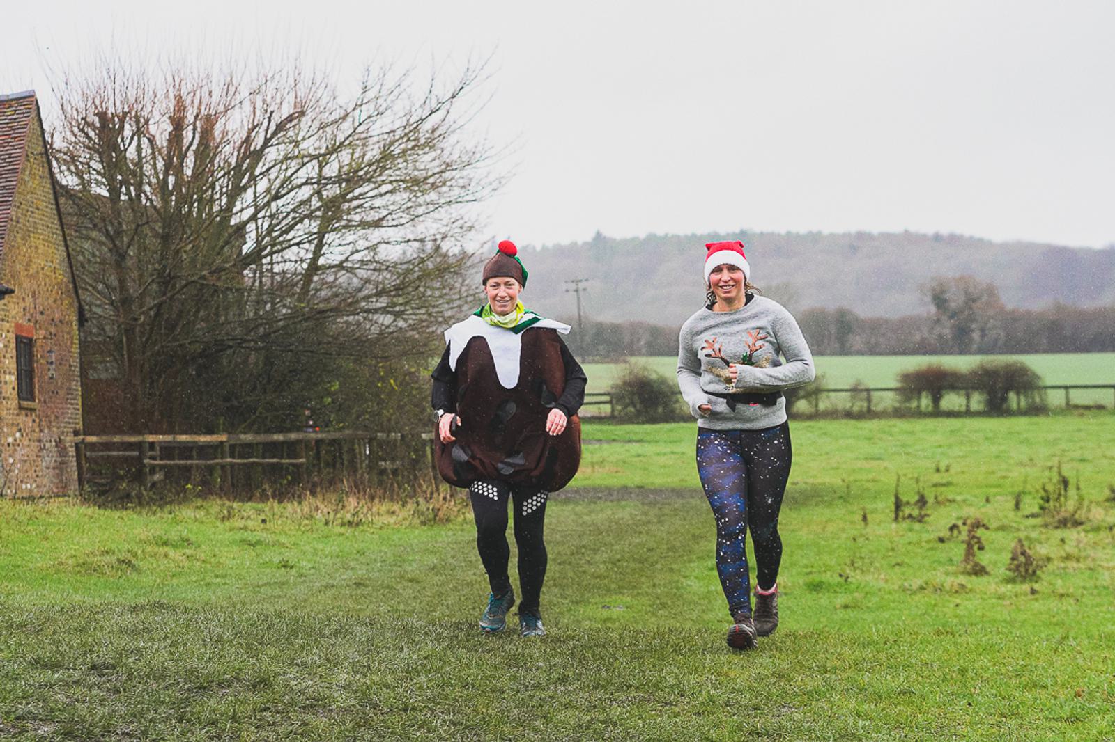 Two people dressed in festive attire run on a grassy path near a brick building and a tree. One person wears a Christmas pudding costume, and the other wears a Santa hat and grey sweater with a reindeer design. The landscape features open fields and rolling hills.