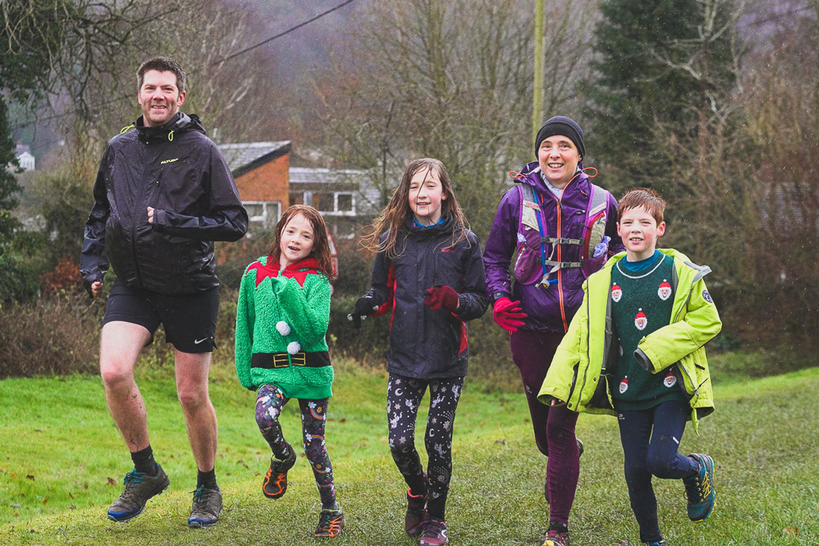 A group of five people, two adults, and three children, are running on a grassy path. They are dressed in colorful winter running attire. The background features trees and a hint of buildings, suggesting they are in a park or suburban area. Everyone is smiling and enjoying the run.