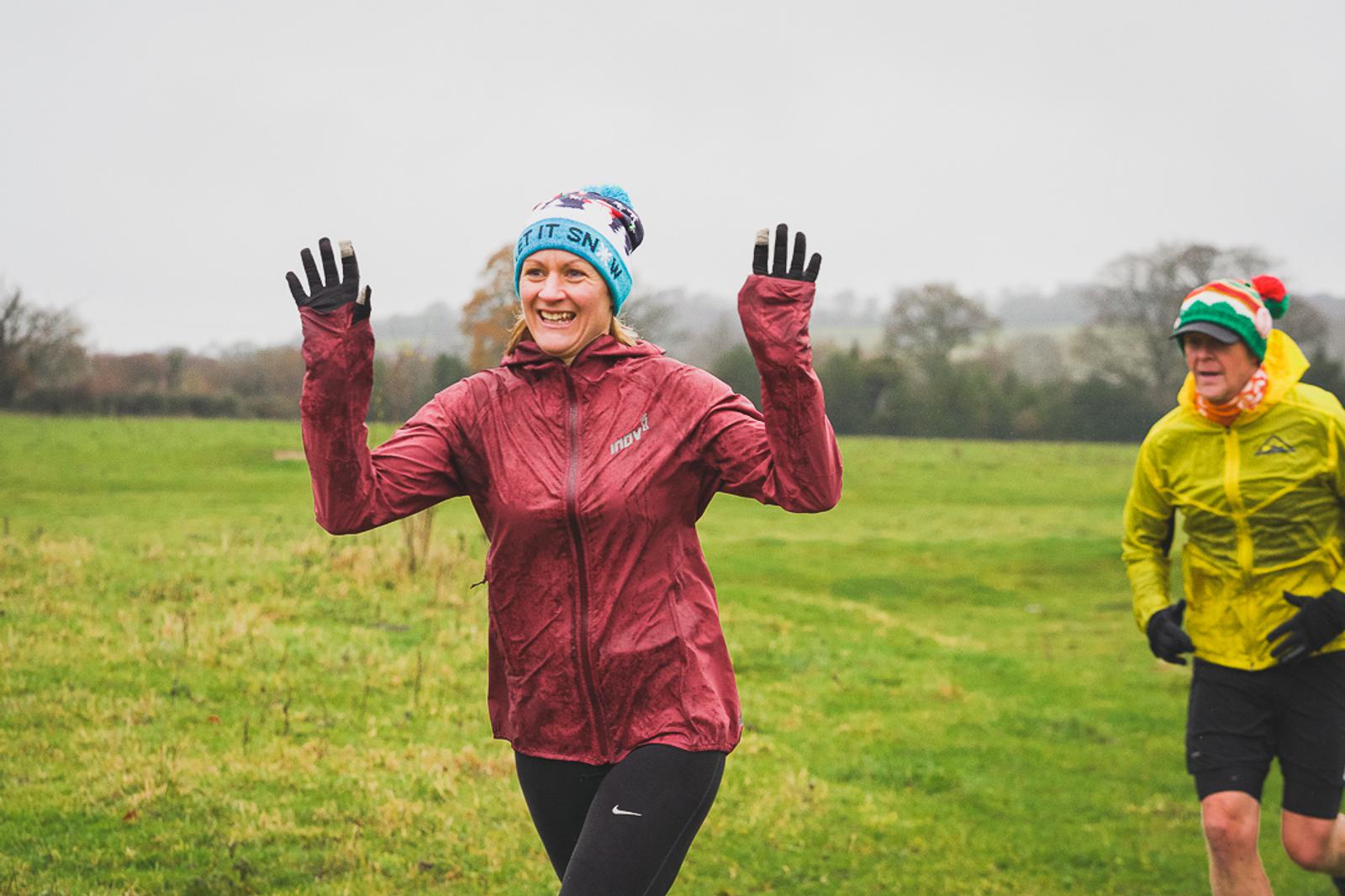 A person wearing a red jacket and a colorful beanie is running outdoors on a grassy field, smiling and raising their hands in the air. Another person in a yellow jacket and festive hat runs behind them. The sky is overcast, suggesting cool weather.