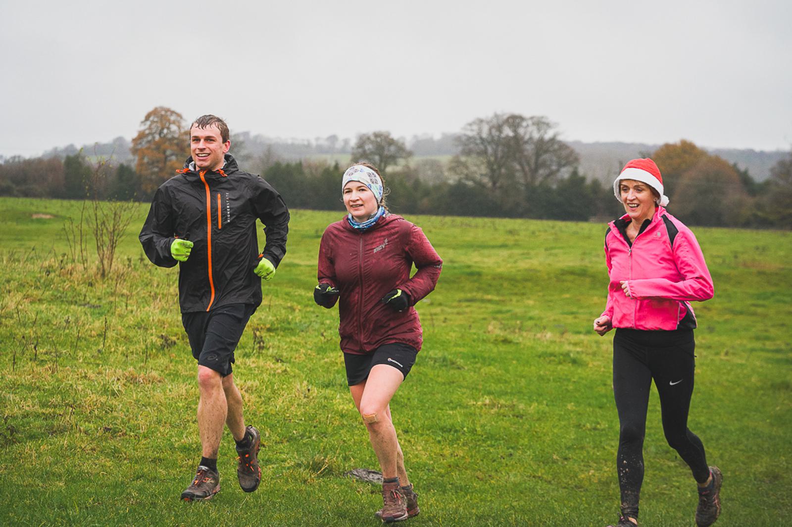 Three people are running on a grassy field under an overcast sky. They are wearing jackets and shorts, with one in a pink jacket and festive Santa hat, another in a brown jacket and beanie, and the third in a black and orange jacket. Trees are visible in the background.