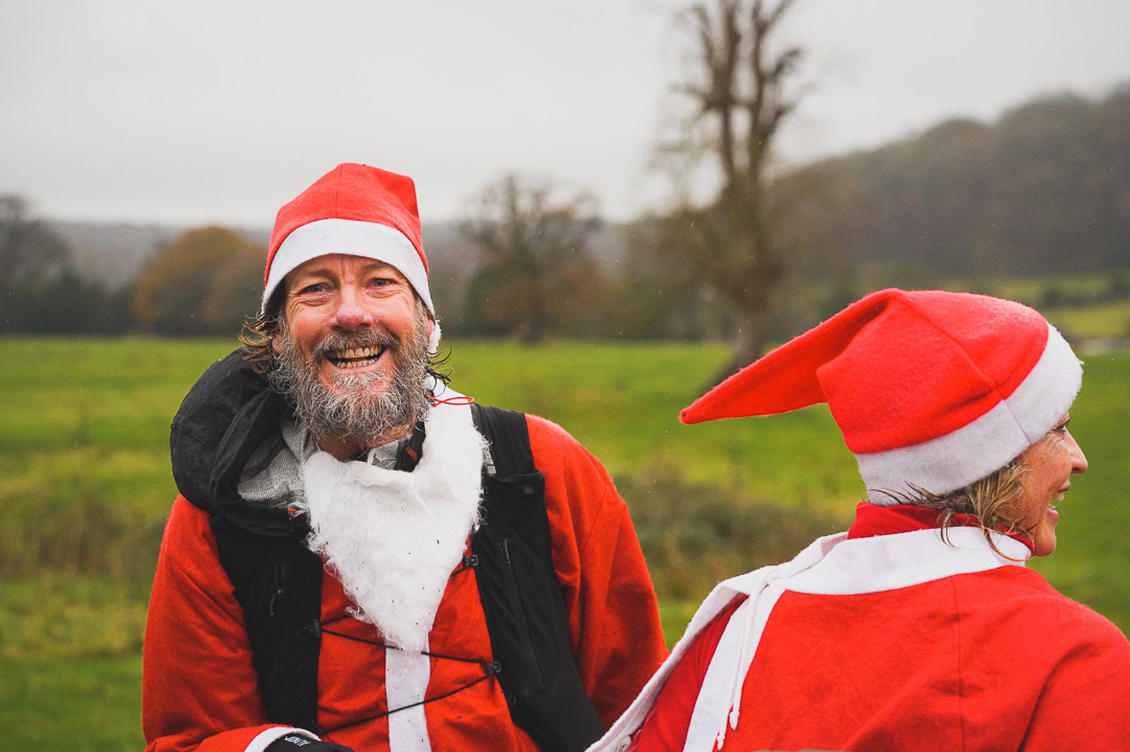 Two people dressed in Santa Claus costumes stand outdoors. The person on the left is smiling with a long beard, while the person on the right, with their back to the camera, wears a Santa hat. They are surrounded by a green field and trees with a cloudy sky above.