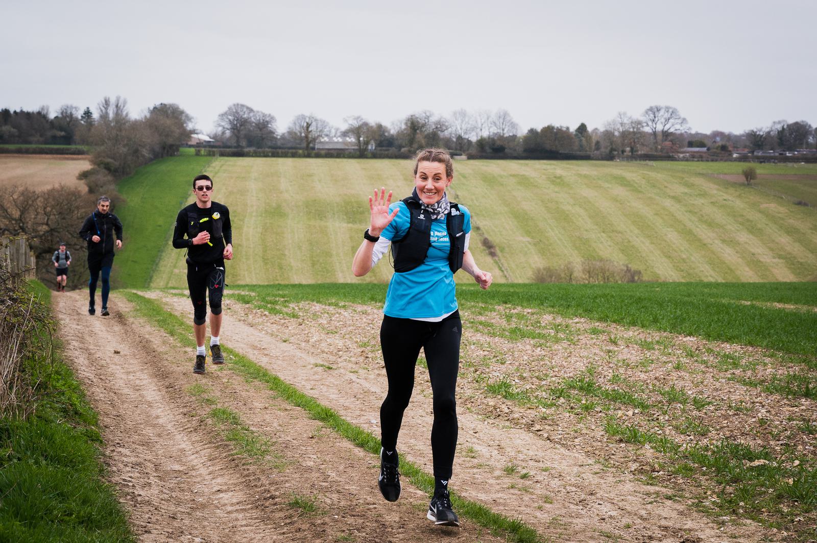 A woman in athletic wear waves and smiles while running on a dirt path through a green countryside. Two other runners follow behind her, with one wearing sunglasses. Trees and fields are visible in the background under an overcast sky.