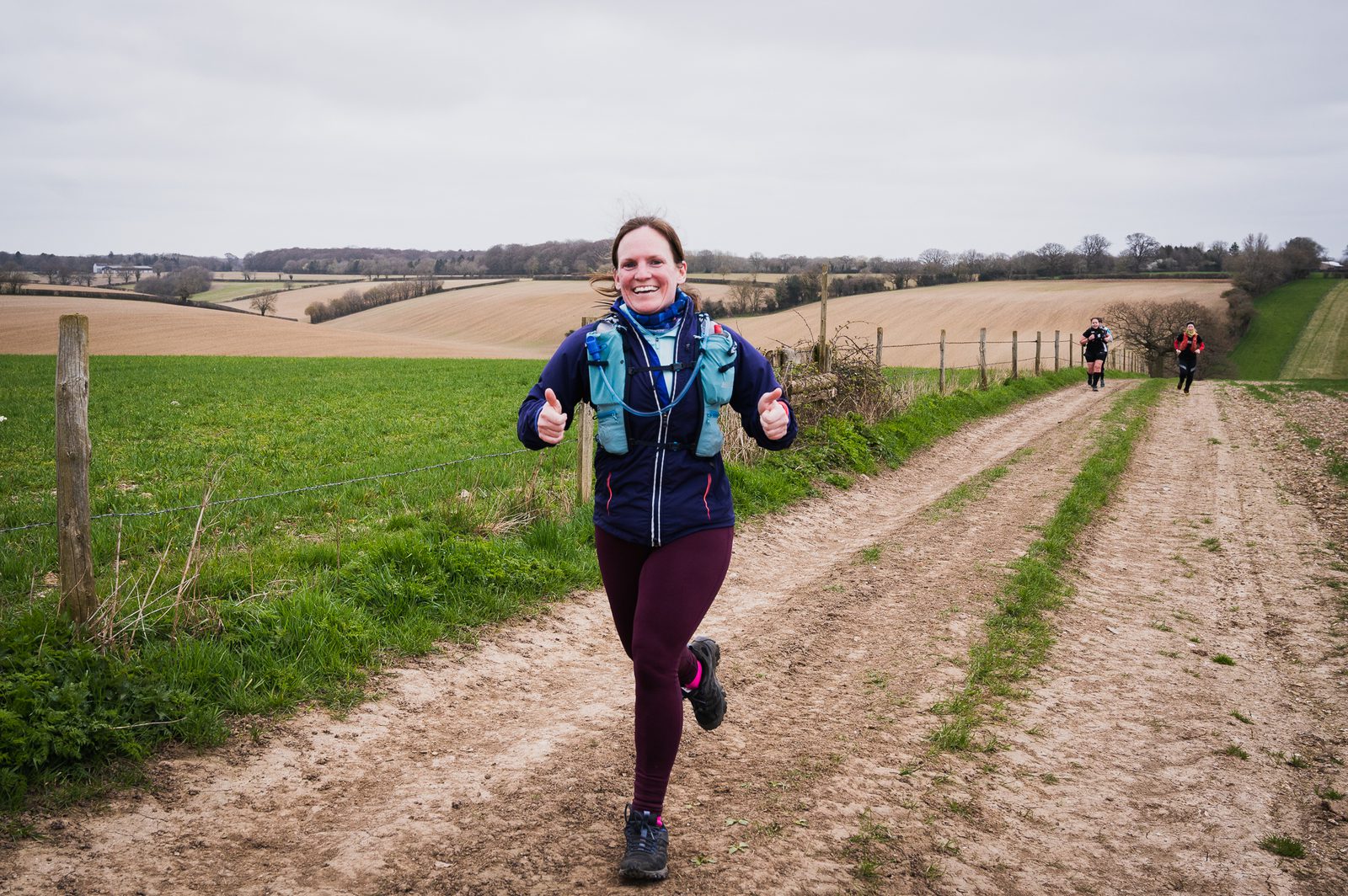 A woman is running along a dirt path through open fields, smiling and giving two thumbs up. She is wearing a blue jacket and maroon leggings, with a hydration pack on her back. The sky is overcast and other runners are visible in the background.