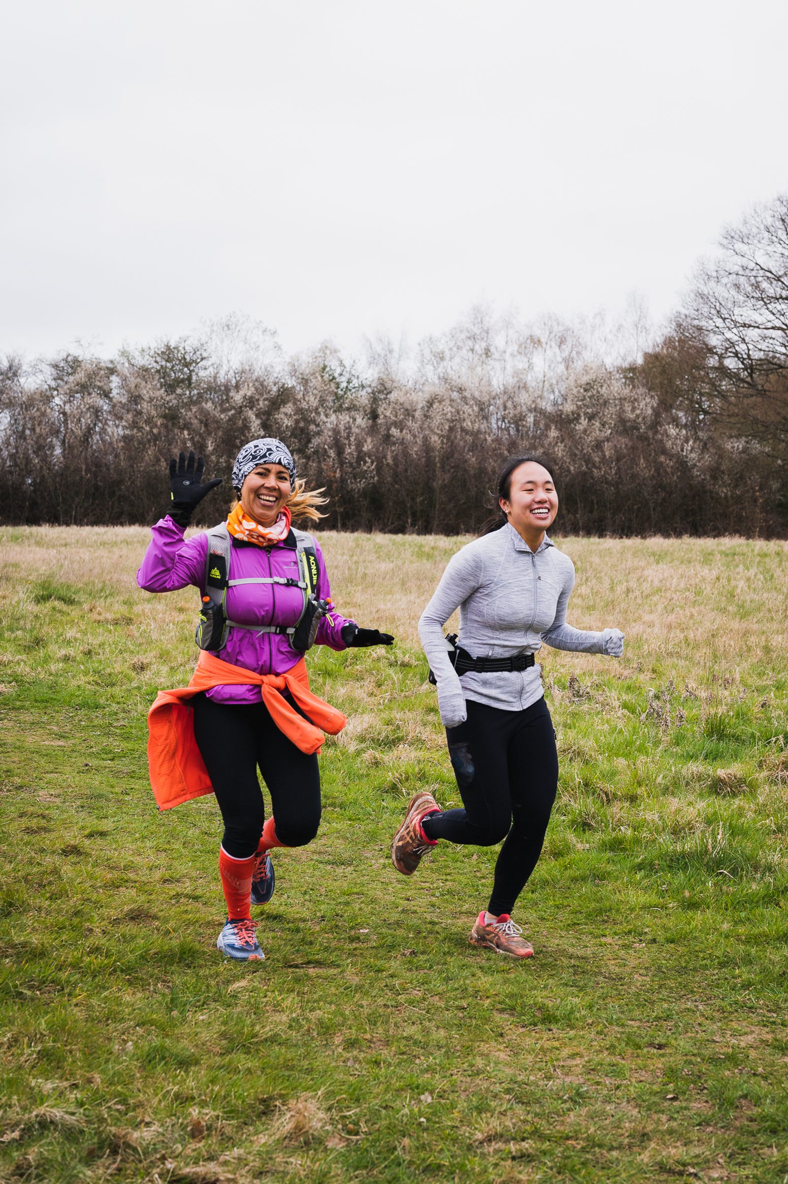 Two people jog on a grassy path in an open field surrounded by trees. The person on the left wears a purple jacket, orange scarf, winter cap, and gloves while waving. The person on the right wears a gray zip-up and black pants. Both are smiling and appear joyful.
