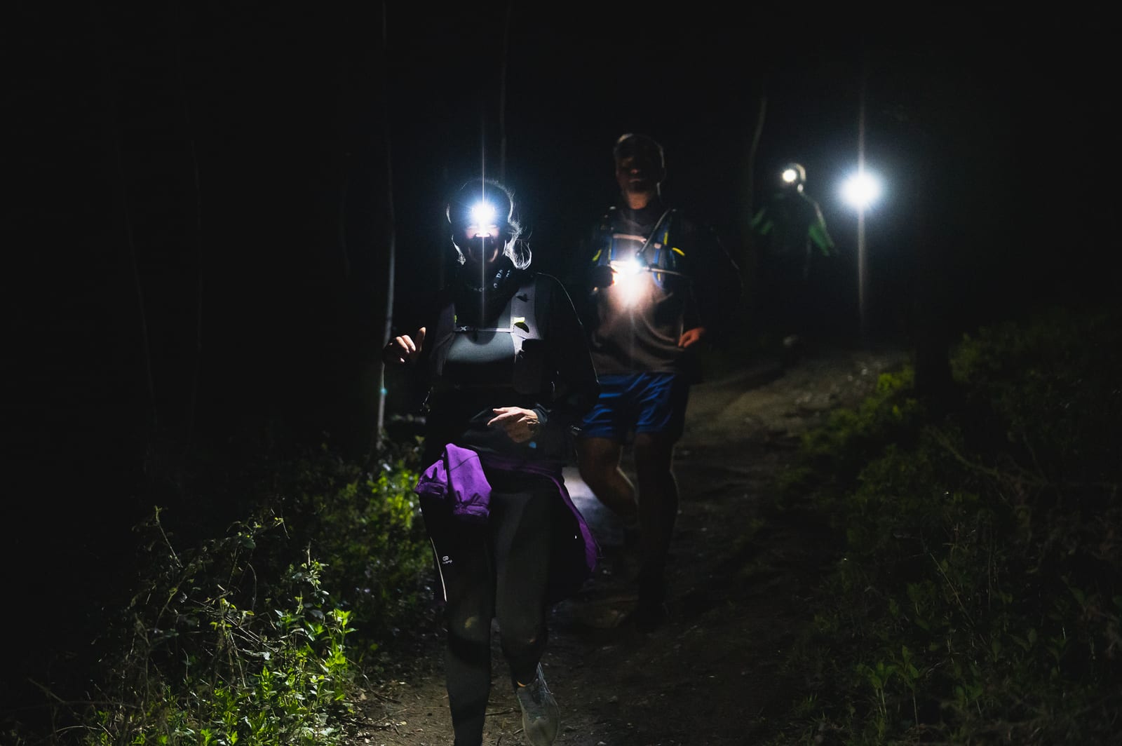 Three people wearing headlamps run along a dark forest trail at night. Their lamps cast bright beams of light, illuminating their paths. The lead runner wears a purple jacket tied around their waist. Sparse vegetation borders the trail.