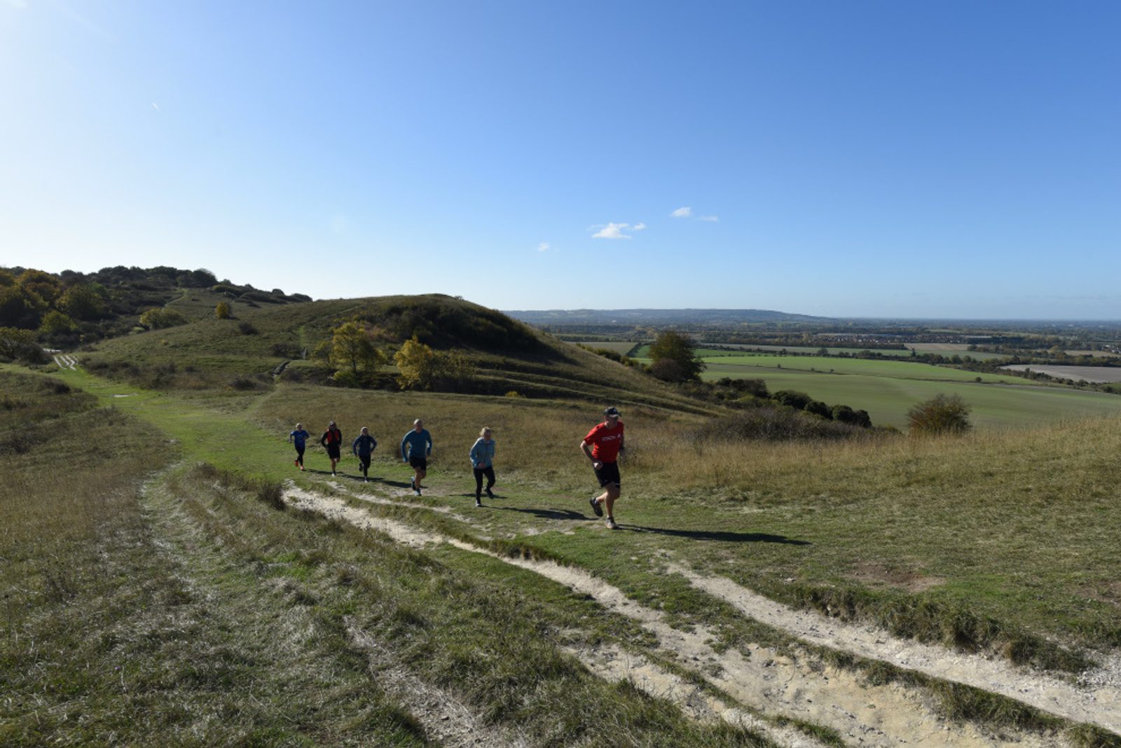 A group of six people are running uphill on a dirt path through a grassy landscape with gentle hills. They are spread out in a line, with one person in a red shirt leading the way. The sky is clear and blue, and the background features fields and distant horizons.