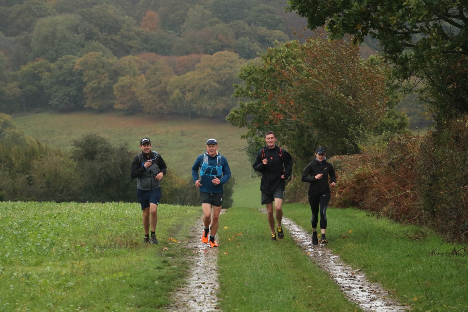 Four people are jogging on a muddy trail surrounded by lush greenery and trees. They wear athletic gear and appear focused. The background features a forested area with a mix of green and autumn-hued foliage. The scene is slightly overcast.