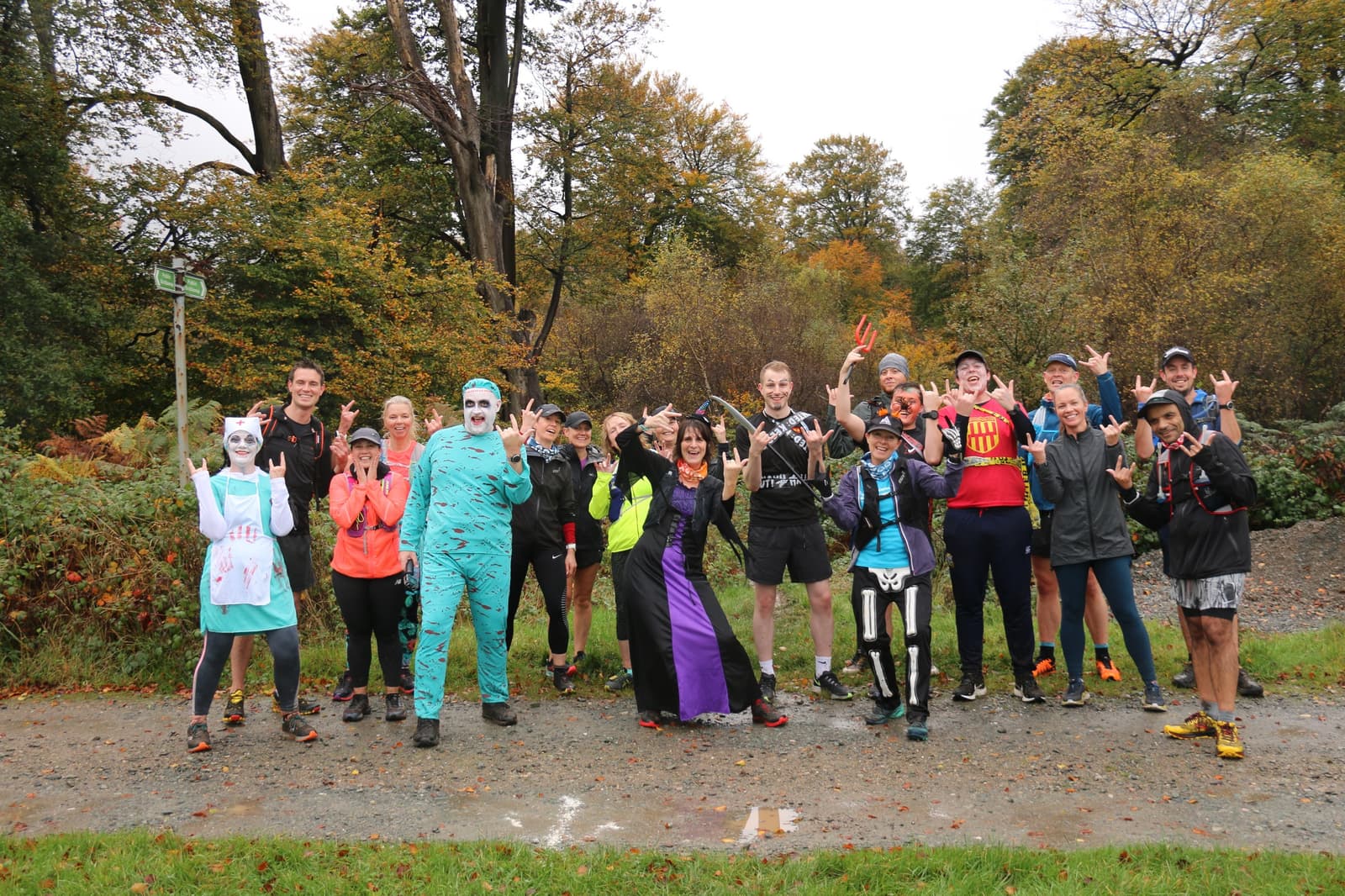 A group of people dressed in various Halloween costumes are standing outdoors on a gravel path surrounded by autumn trees. Some are making playful hand gestures and smiling at the camera. The group is a mix of men and women, all appearing to have fun.