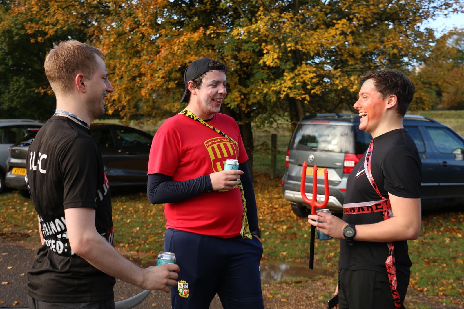 Three people stand outdoors, smiling and talking. One wears a black shirt, another in a red shirt, and the third in a black shirt with a red headband, holding a trident. They are in a parking lot with parked cars and trees with autumn foliage in the background.