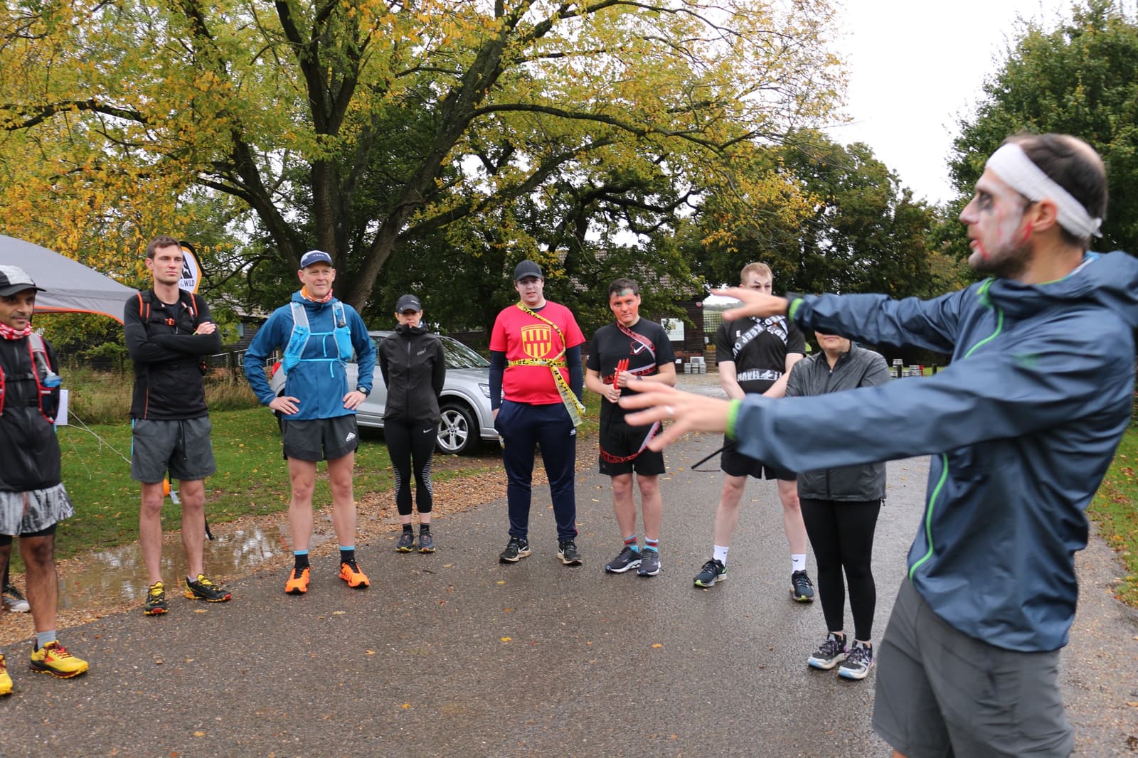 A group of runners in athletic gear gather around a person in a blue jacket and white headband who is giving instructions. The scene is set outdoors with trees showing autumn colors. Some participants are attentively listening while others look relaxed.