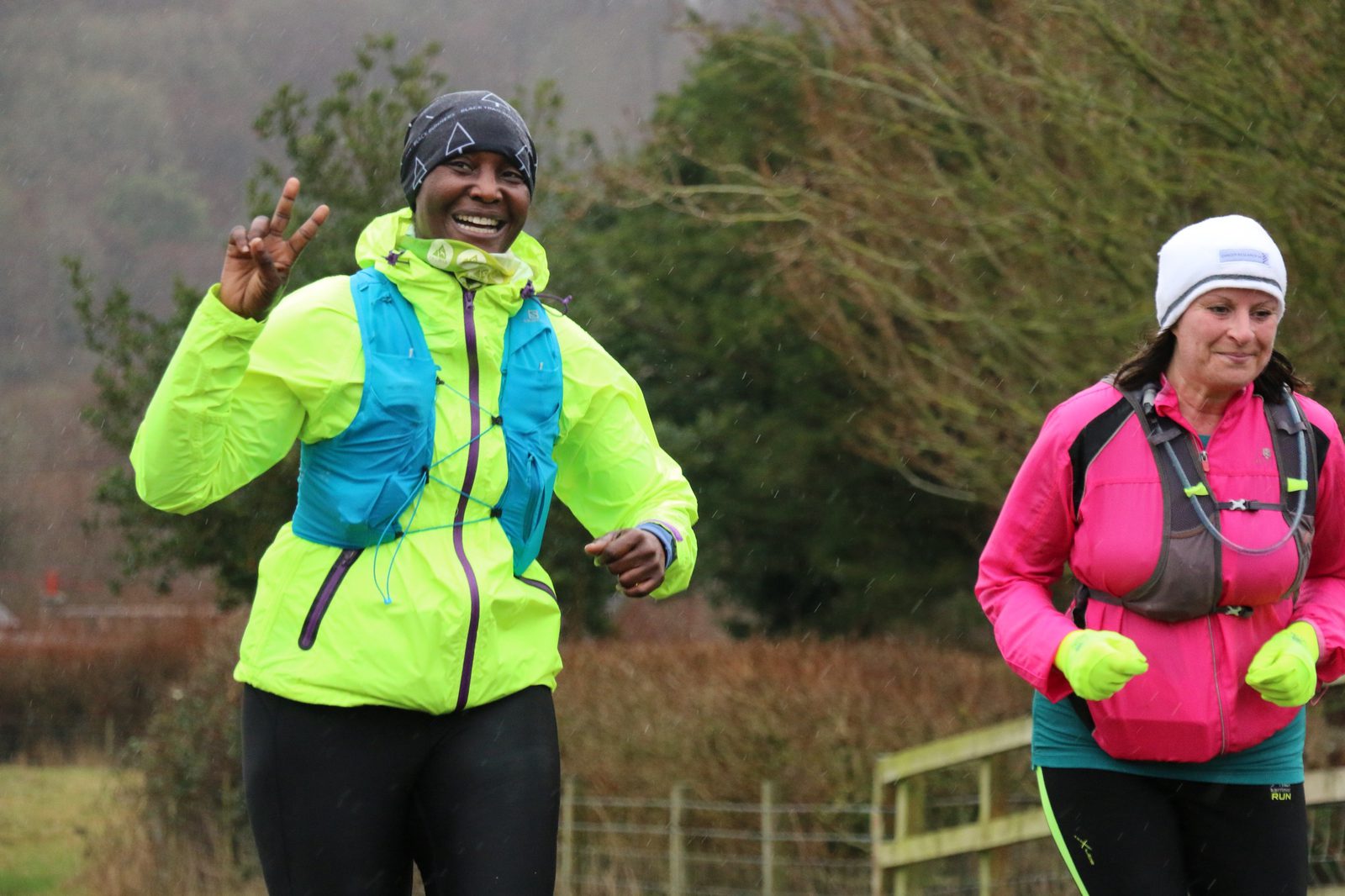 Two people running outside in a park. The person on the left is smiling and making a peace sign with their hand, wearing a neon yellow jacket and blue vest. The person on the right is wearing a pink jacket and white beanie. Both appear to be enjoying their run.