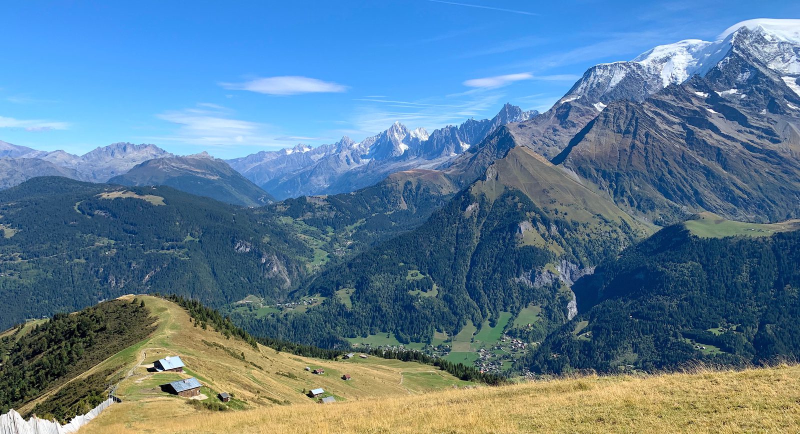 A panoramic view of a mountainous landscape under a clear blue sky. Snow-capped peaks tower in the background, while lush green valleys and scattered rustic cabins dot the foreground hills. A walking path meanders through the grassy slope in the lower left corner.