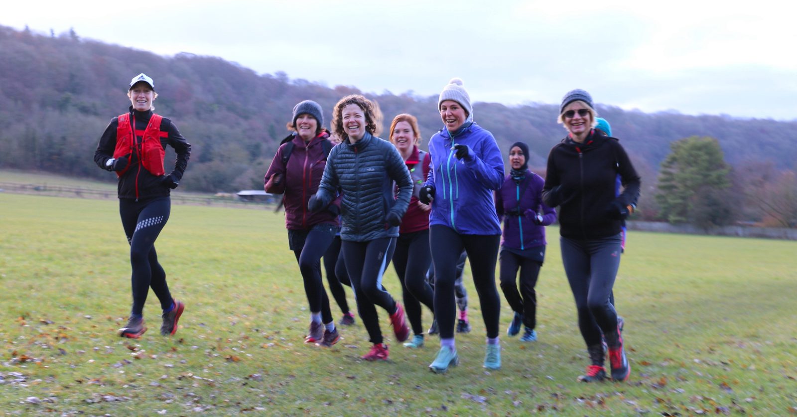 A group of people dressed in winter running gear jogs together in a grassy field surrounded by trees. They are smiling and appear to be enjoying the activity. The sky is overcast, and the participants are wearing jackets, hats, and gloves to stay warm.
