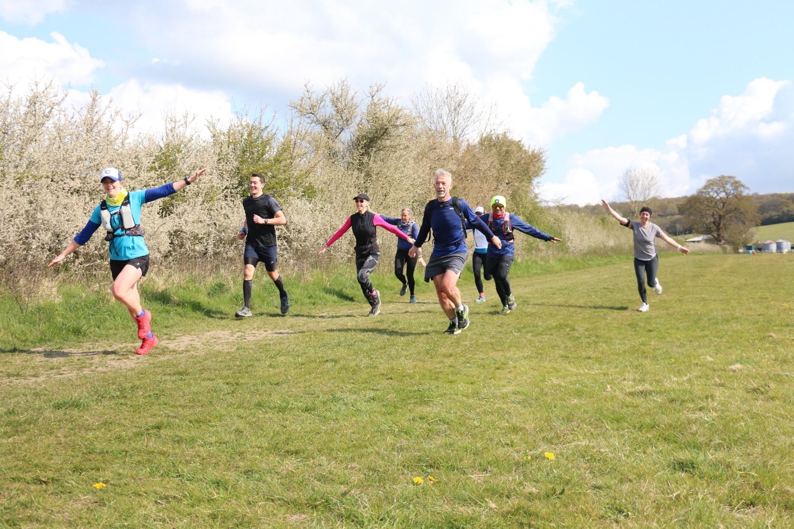 A group of six people holding hands and running on a grassy path near some bushes. They appear joyful and energetic under a partly cloudy sky. One person is ahead leading the group, while the others follow closely behind, smiling and with arms outstretched.