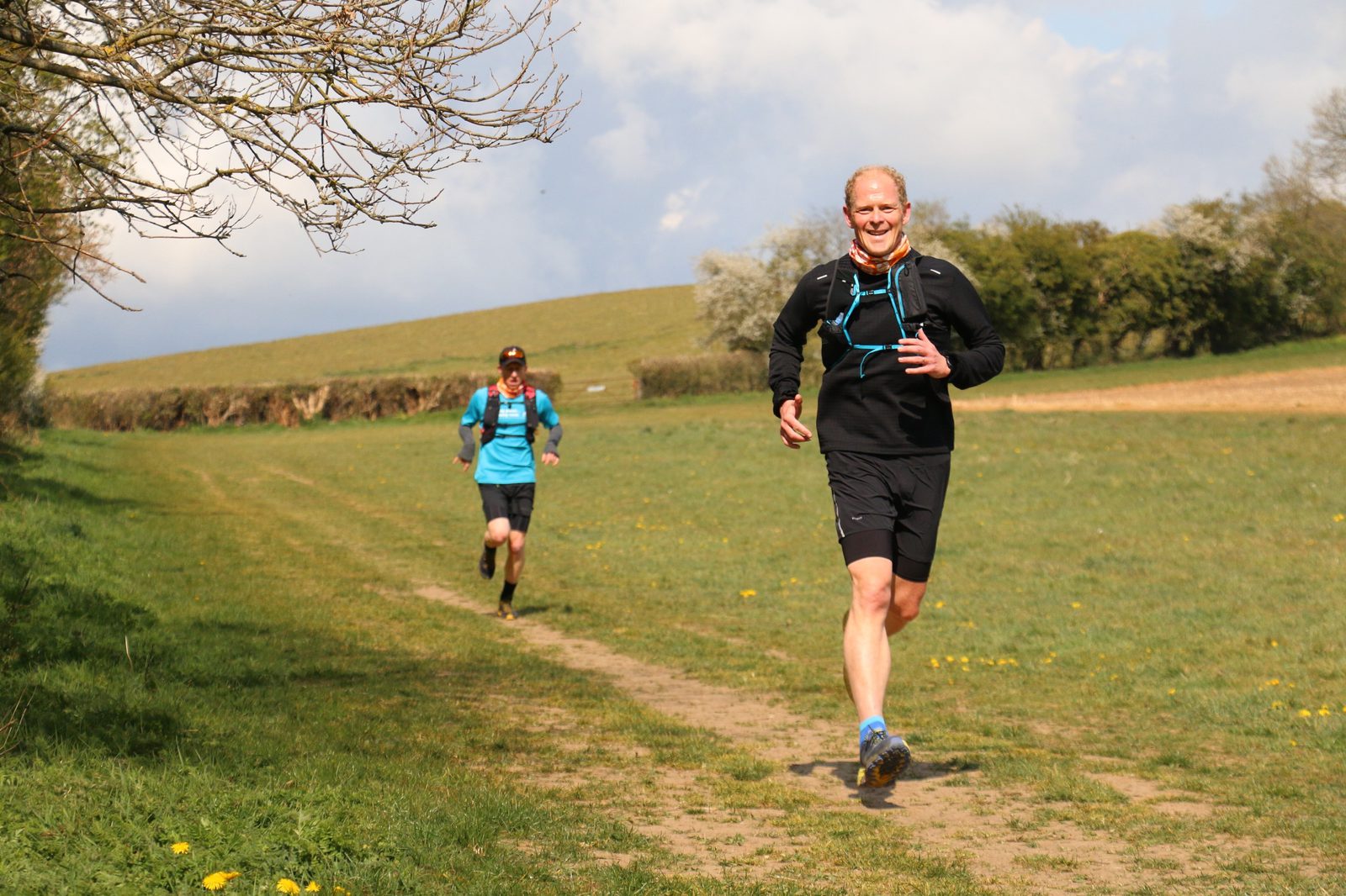 Two people running on a grassy trail through a rural area. The closer runner is smiling, wearing black shorts and a long-sleeve top with blue accents. The second runner is further back, wearing a blue shirt and dark shorts. The landscape includes trees, a hedge, and open fields.