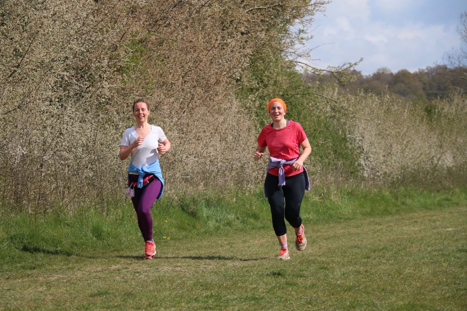 Two women are jogging outdoors on a grassy path beside blooming bushes with light pink flowers. They both smile as they run, wearing athletic clothes; one wears a red shirt and headband, while the other wears a white shirt. The sky in the background is partly cloudy.