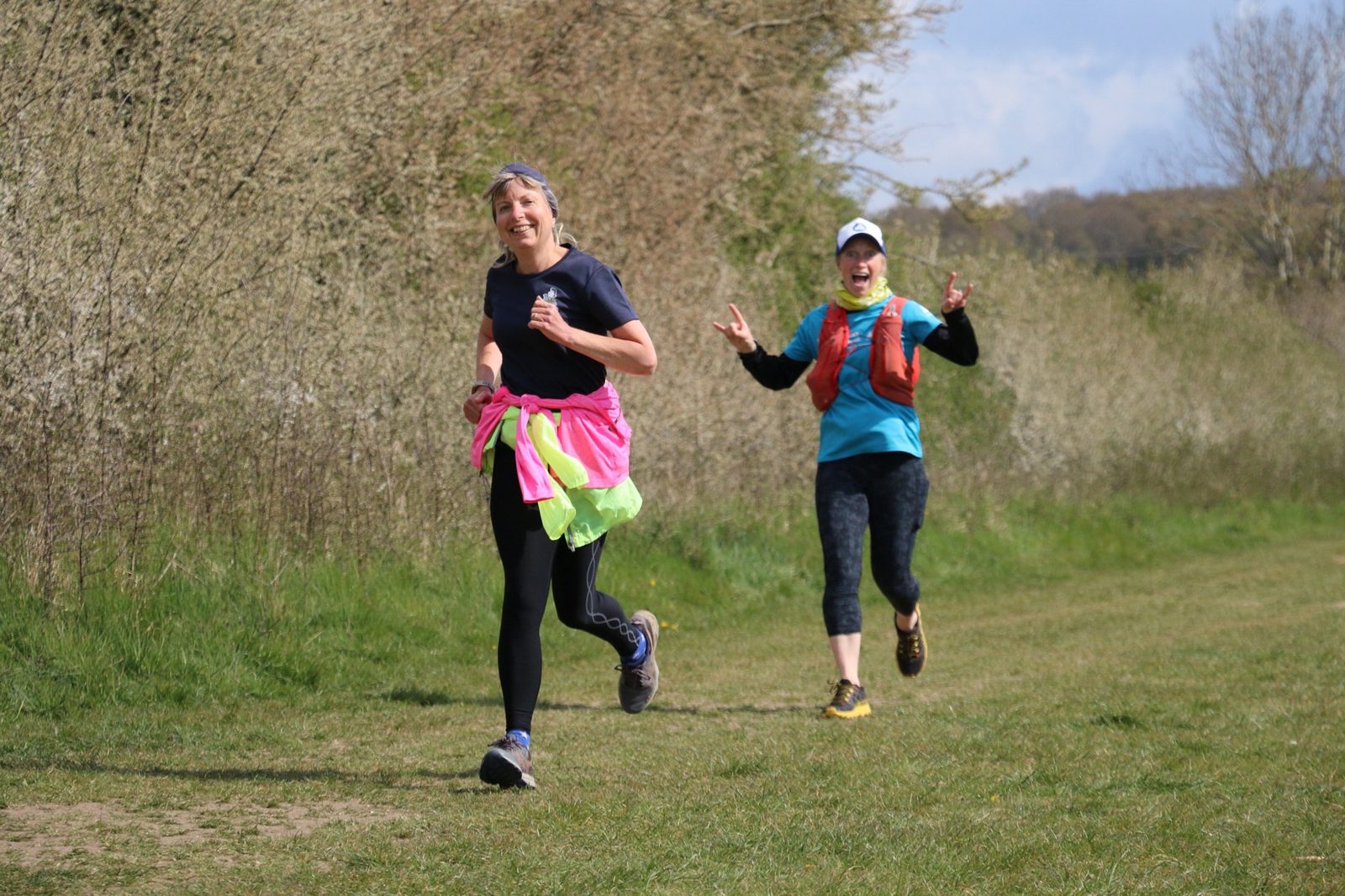 Two women are running on a grassy trail with trees and shrubs in the background. The woman in the front is smiling, wearing a black outfit with a colorful jacket tied around her waist. The woman behind her is also smiling, making a peace sign with both hands.