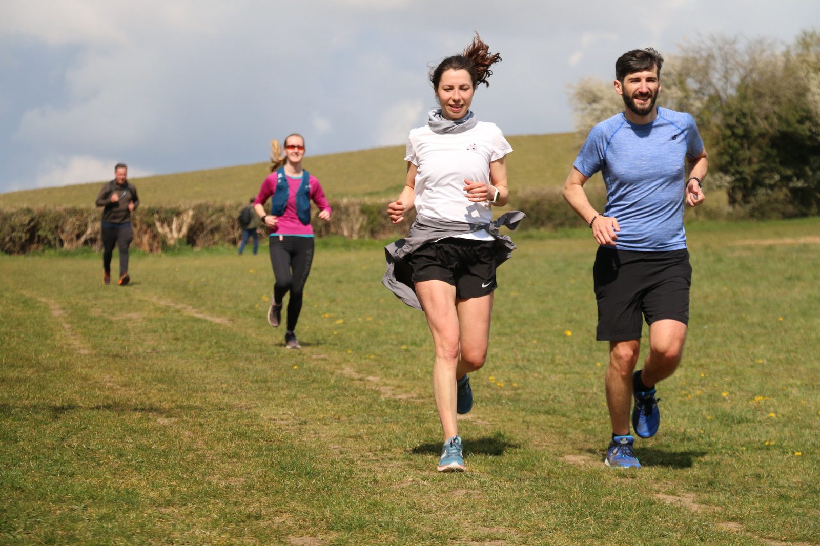 A group of four people are running outdoors on a grassy trail. The two runners in the front, a man in a blue shirt and a woman in a white shirt, are smiling. Two other runners are in the background, one wearing pink sleeves and another in all black. It's a sunny day.