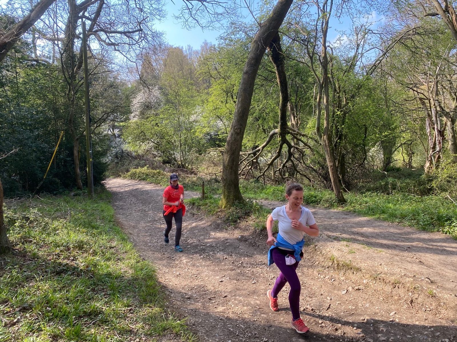 Two women are running on a dirt path in a wooded area with green trees and bushes around them. One runner, wearing a red shirt and black pants, follows closely behind another runner in a white shirt and purple pants. Sunlight filters through the trees.