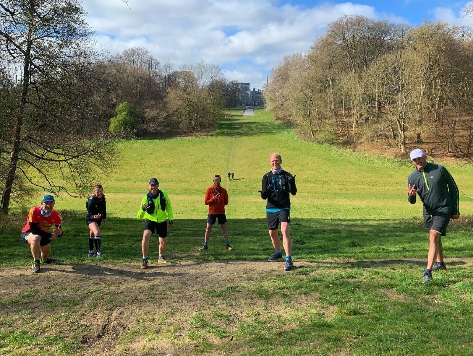 A group of six people dressed in athletic clothing are posing outdoors on a grassy field. They are all smiling and making various playful gestures. Behind them is a wooded area, and in the distance, a building can be seen atop a hill. It is a bright, sunny day.