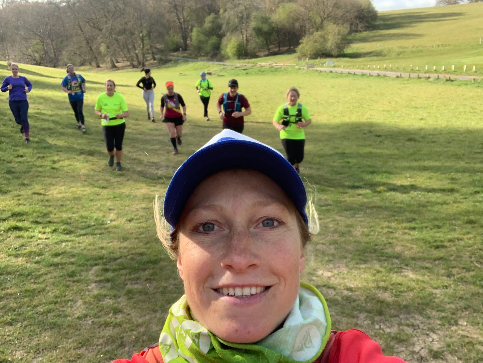 A person wearing a blue cap and green scarf takes a selfie while smiling. In the background, a group of runners are jogging on a grassy field with trees in the distance under a clear sky. The runners are wearing bright athletic gear.