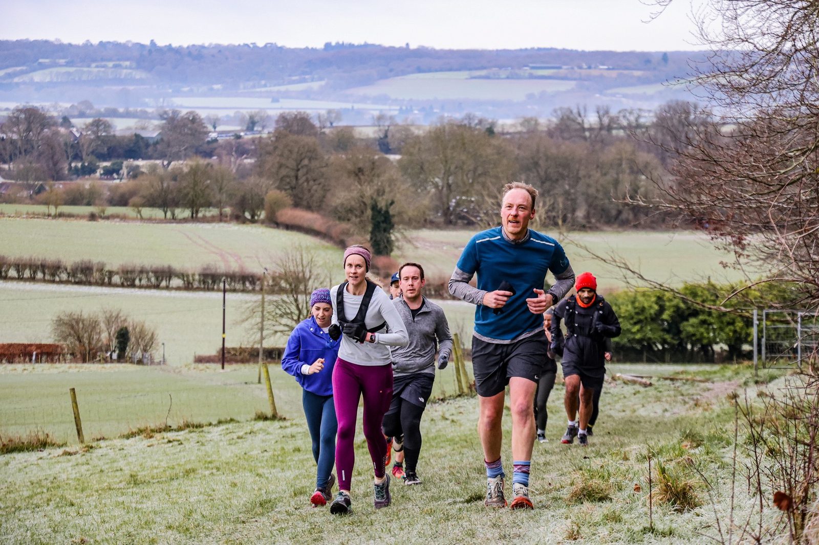 A group of five people jog up a grassy, slightly frosty hill in a rural countryside setting, with rolling fields and trees in the background under a cloudy sky.