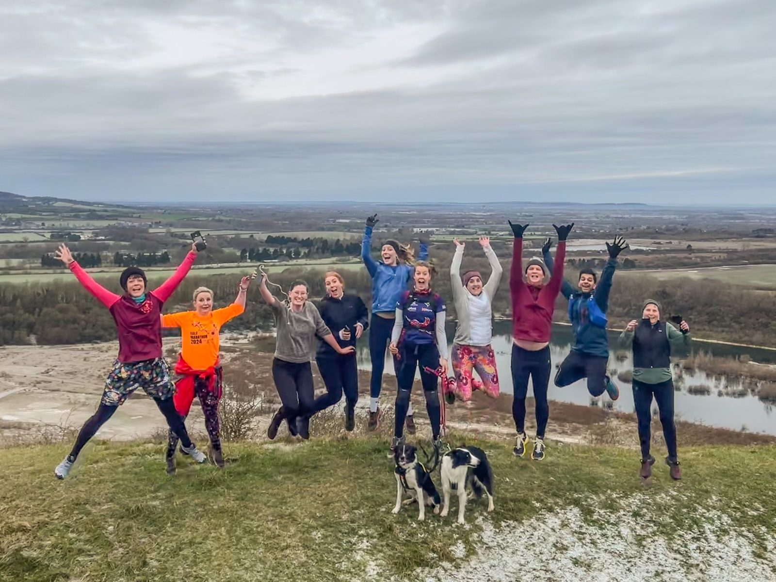 A group of twelve people and two dogs are outdoors on a hill, jumping in the air and smiling. The landscape behind them features fields, water, and a cloudy sky.