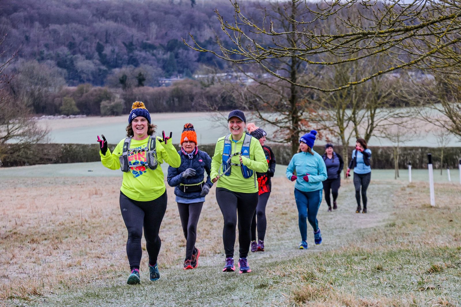 A group of people in colorful winter running gear jog and smile along a frosty, grassy trail in a rural landscape with trees and hills in the background.
