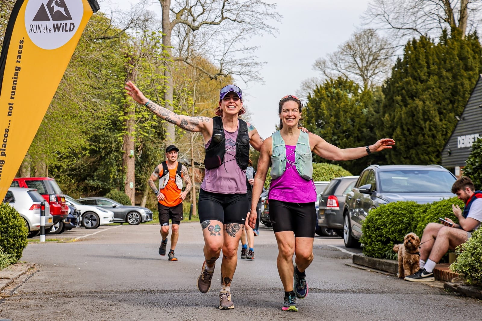 Two smiling women wearing sportswear finish a race together, arms raised in celebration. Other runners and parked cars are in the background. A yellow "Run the Wild" banner is on the left, and a dog sits near spectators on the right.