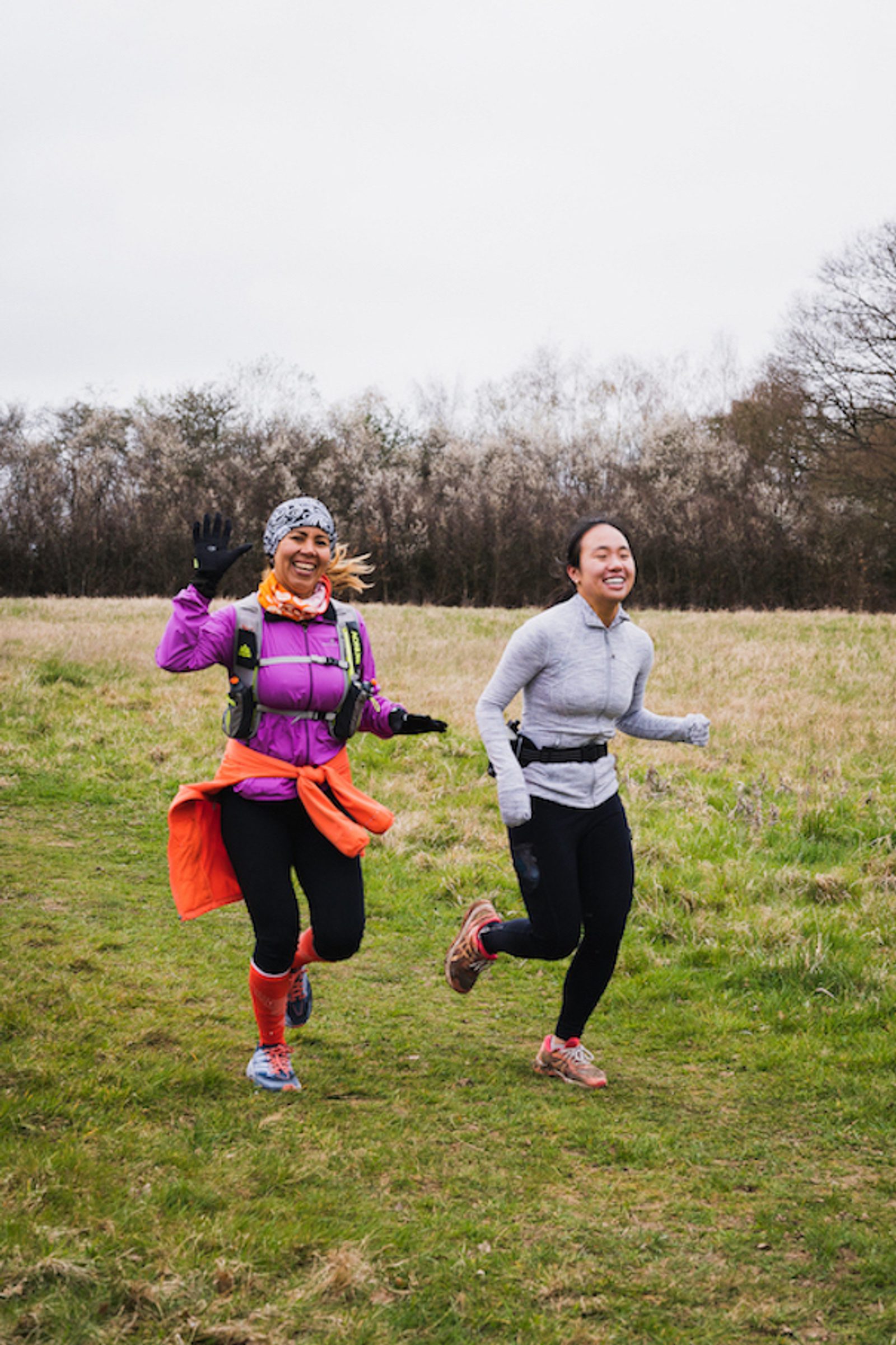 Two women are running on a grassy trail. One is wearing a pink jacket and a headband, waving with her left hand, and the other is in a gray jacket. Both are smiling and seem to be enjoying their run. Trees are visible in the background under an overcast sky.