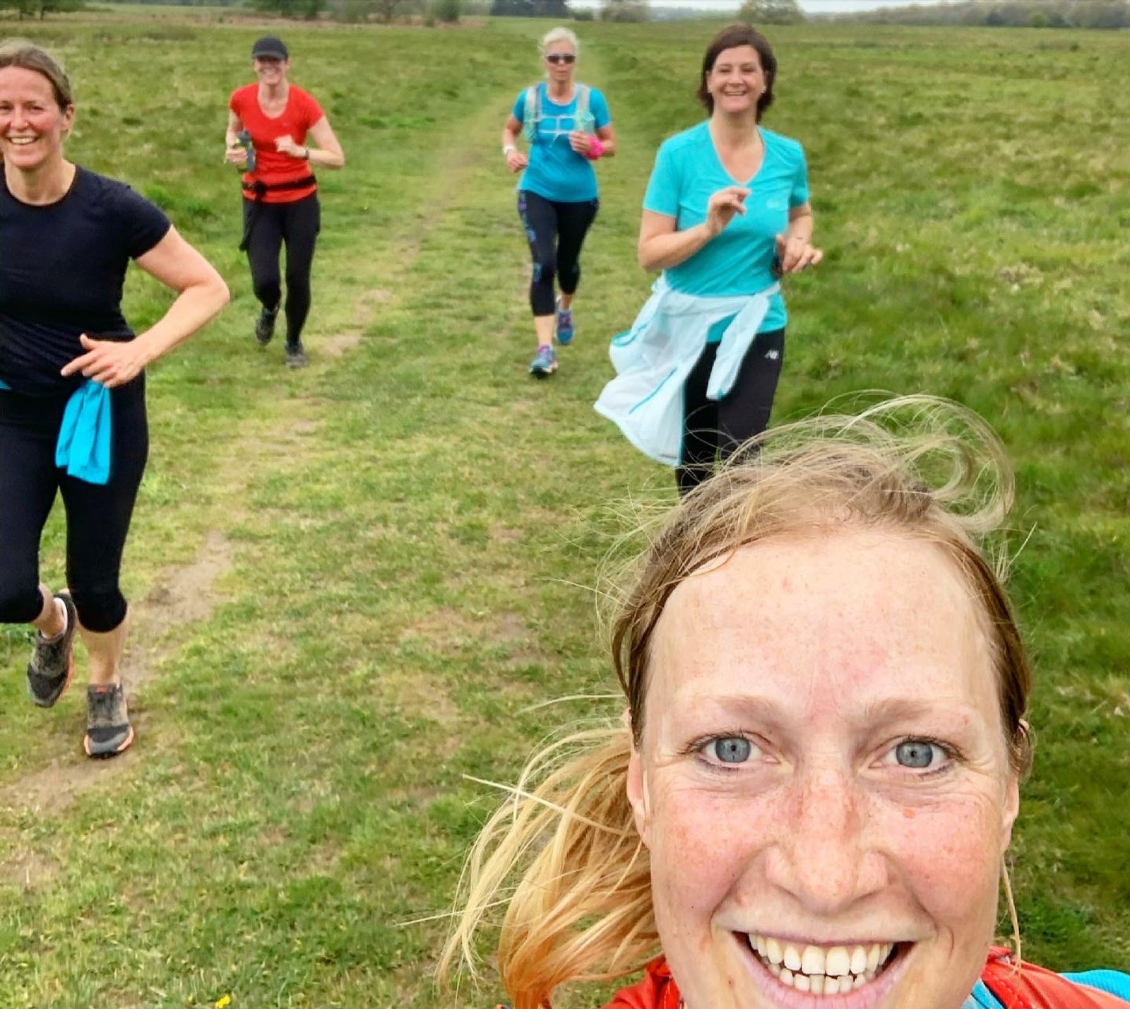 A group of five people are running on a grassy trail. The woman in the foreground is smiling at the camera, while the others run behind her. The sky is overcast, and they appear to be enjoying a run in a natural setting.