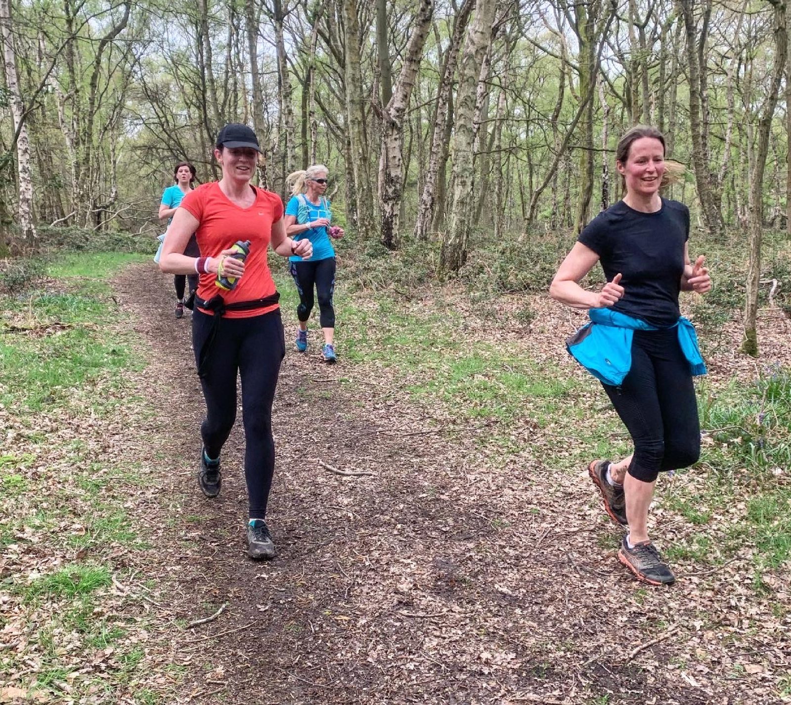 Four people are jogging on a forest trail surrounded by trees and greenery. The two in the foreground are wearing athletic clothing, with one in a red top and black cap, and the other in a black top with a blue jacket around their waist. Everyone seems to be enjoying the run.