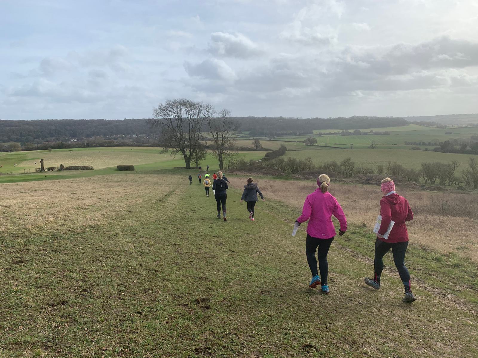 A group of people jogs down a grassy, slightly muddy path in a hilly countryside landscape. They wear bright athletic clothing and run in a line, with expansive fields and scattered trees under a cloudy sky in the background.