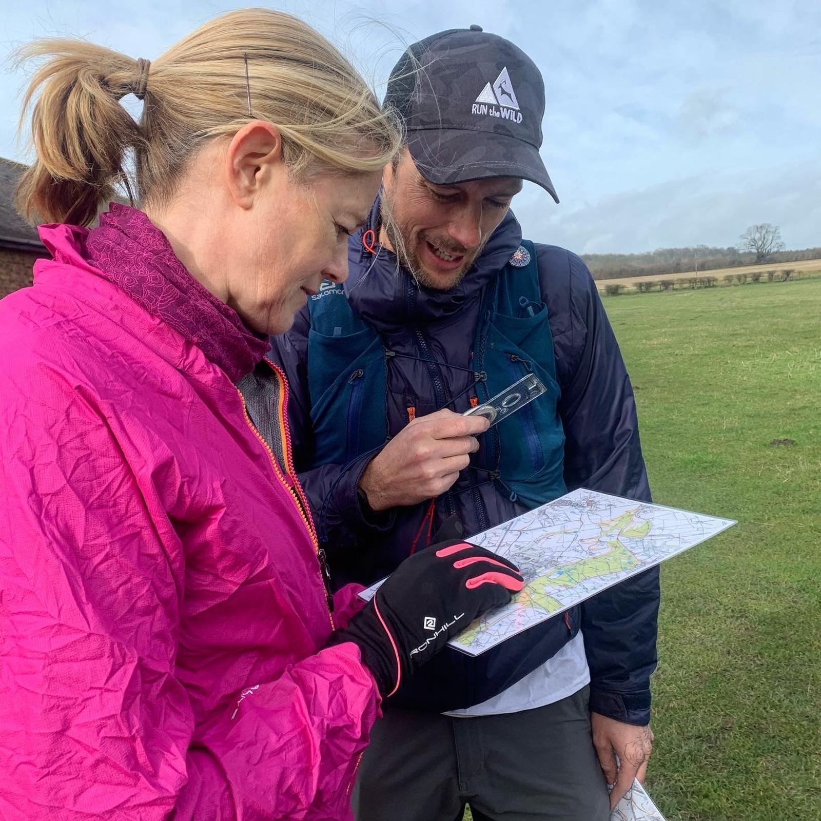 Two individuals are outdoors, engaged in navigating a map. The person on the left wears a bright pink jacket and gloves, while the person on the right, in a hat and dark jacket, looks at the map and holds a compass or phone. They are in an open grassy area with fields in the background.