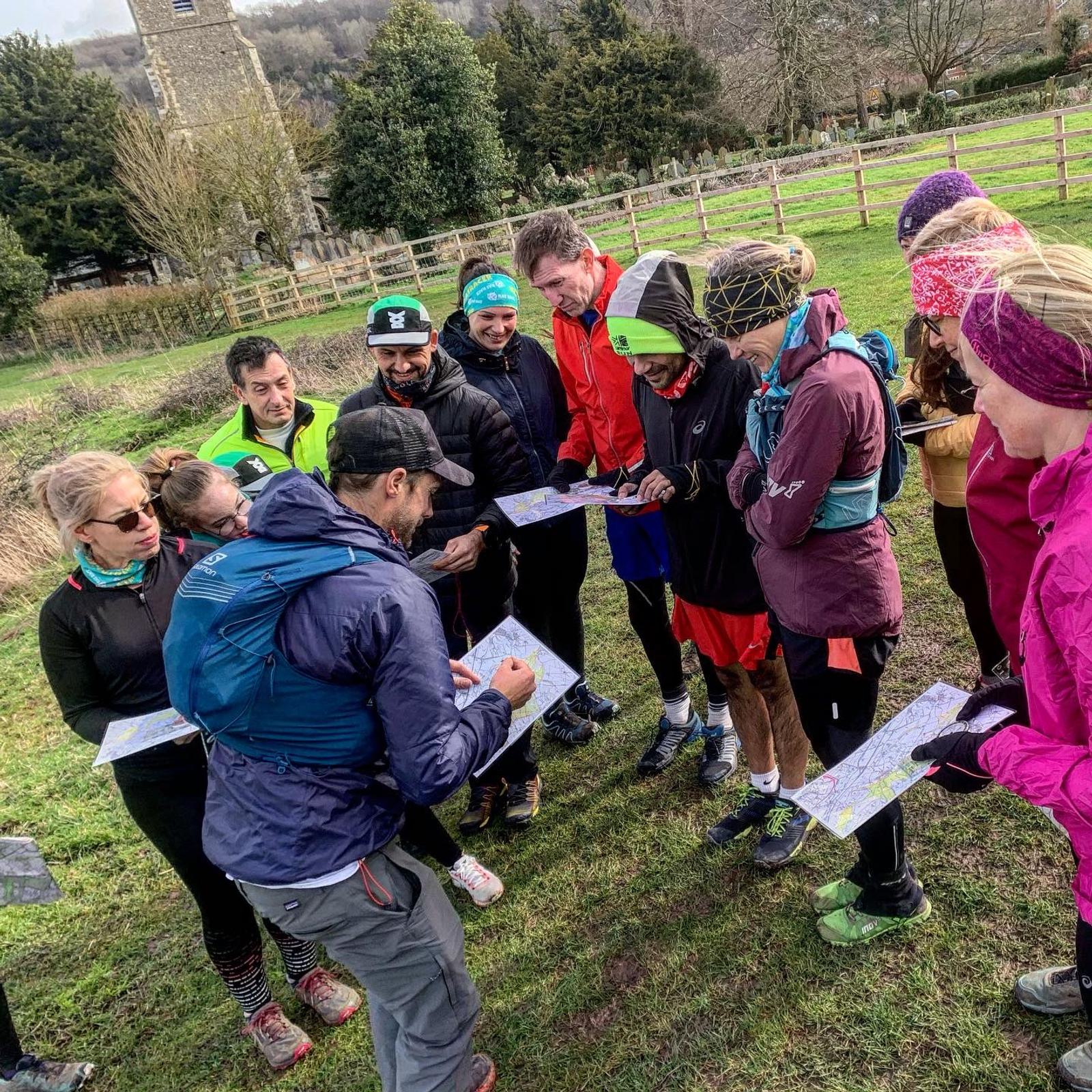 A group of people stand outdoors in a circle, attentively looking at a man pointing at maps. They are dressed in outdoor clothing suitable for hiking or running. Trees, a building, and a fence are visible in the background, suggesting a rural or park setting.