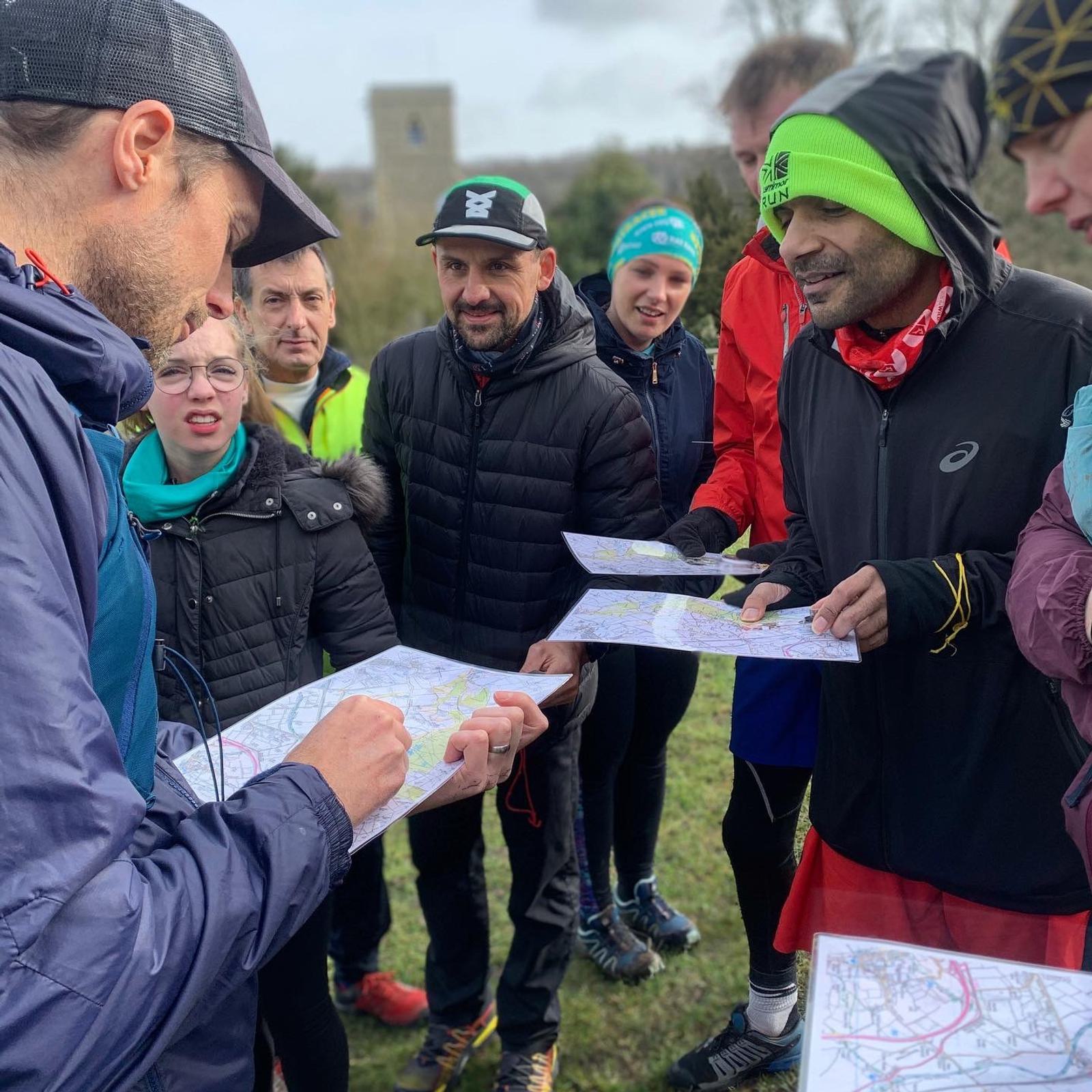 A group of people dressed in athletic gear are gathered outdoors. They are looking intently at maps as one person in a cap and jacket makes notes on a map. The background includes trees and a tall stone structure.