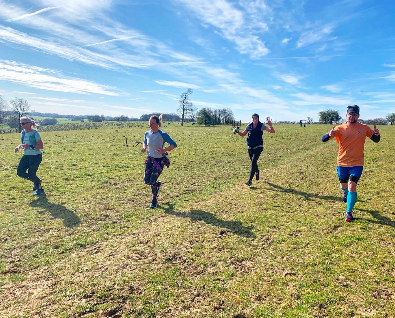 Four people run on a grassy trail in bright sunlight. Three are wearing jackets, and one is in a short sleeve shirt and shorts. The sky is clear blue with streaks of white clouds. They seem to be enjoying the run, with one person giving a thumbs-up.