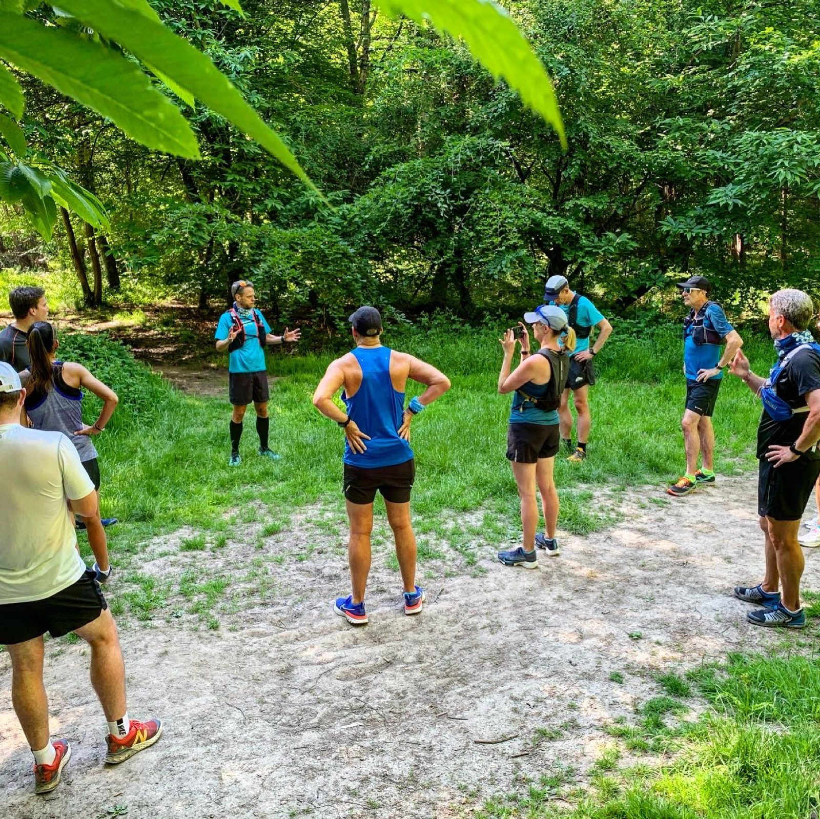 A group of runners gather in a forested area, standing in a loose circle on a small dirt path. One person appears to be leading or giving instructions, while others listen attentively, with some hands on hips or adjusting their gear. The setting is lush and green.