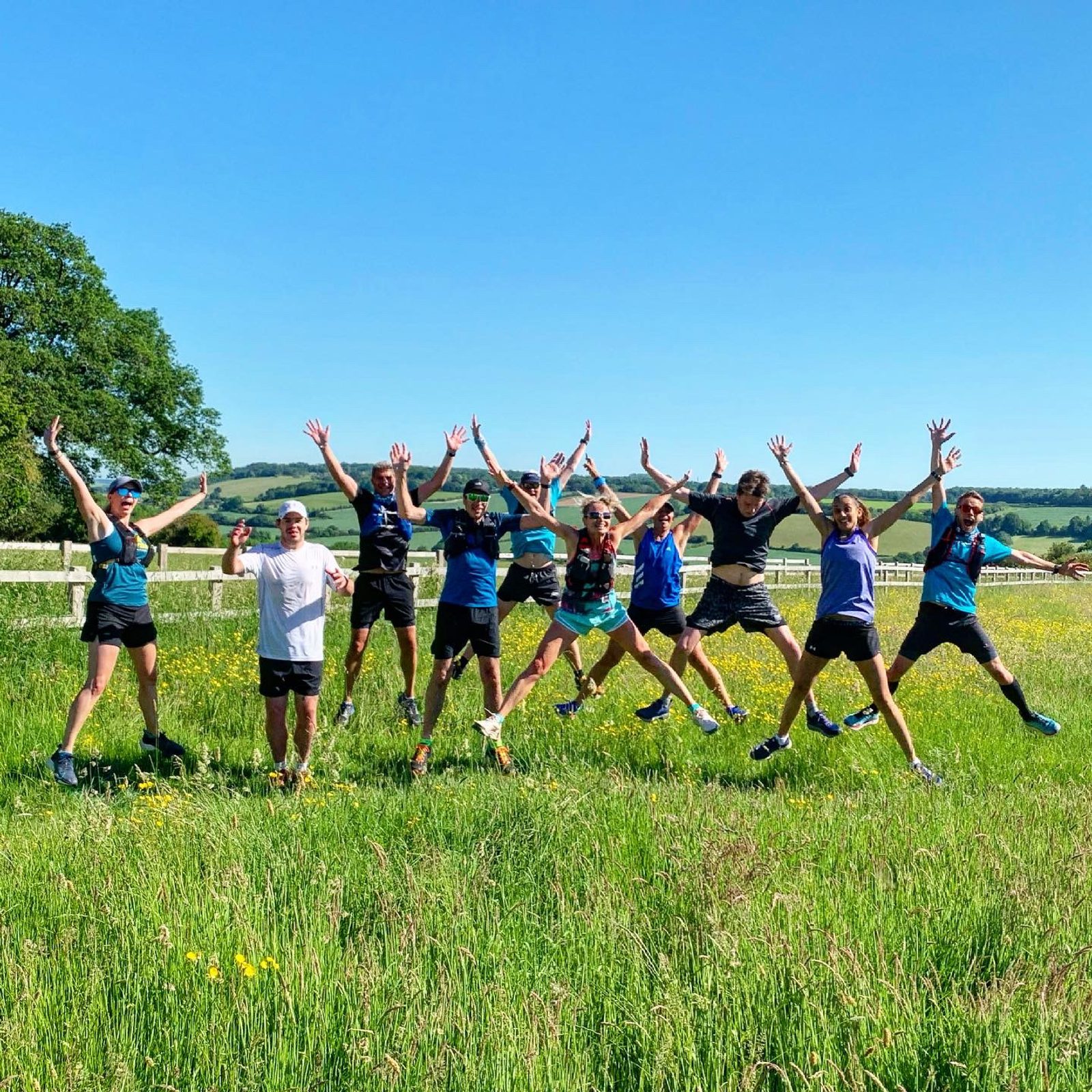 A group of ten people is jumping in the air with their arms raised, smiling and posing for the camera. They are outdoors in a grassy field on a sunny day with a clear blue sky. Trees and distant hills are visible in the background.