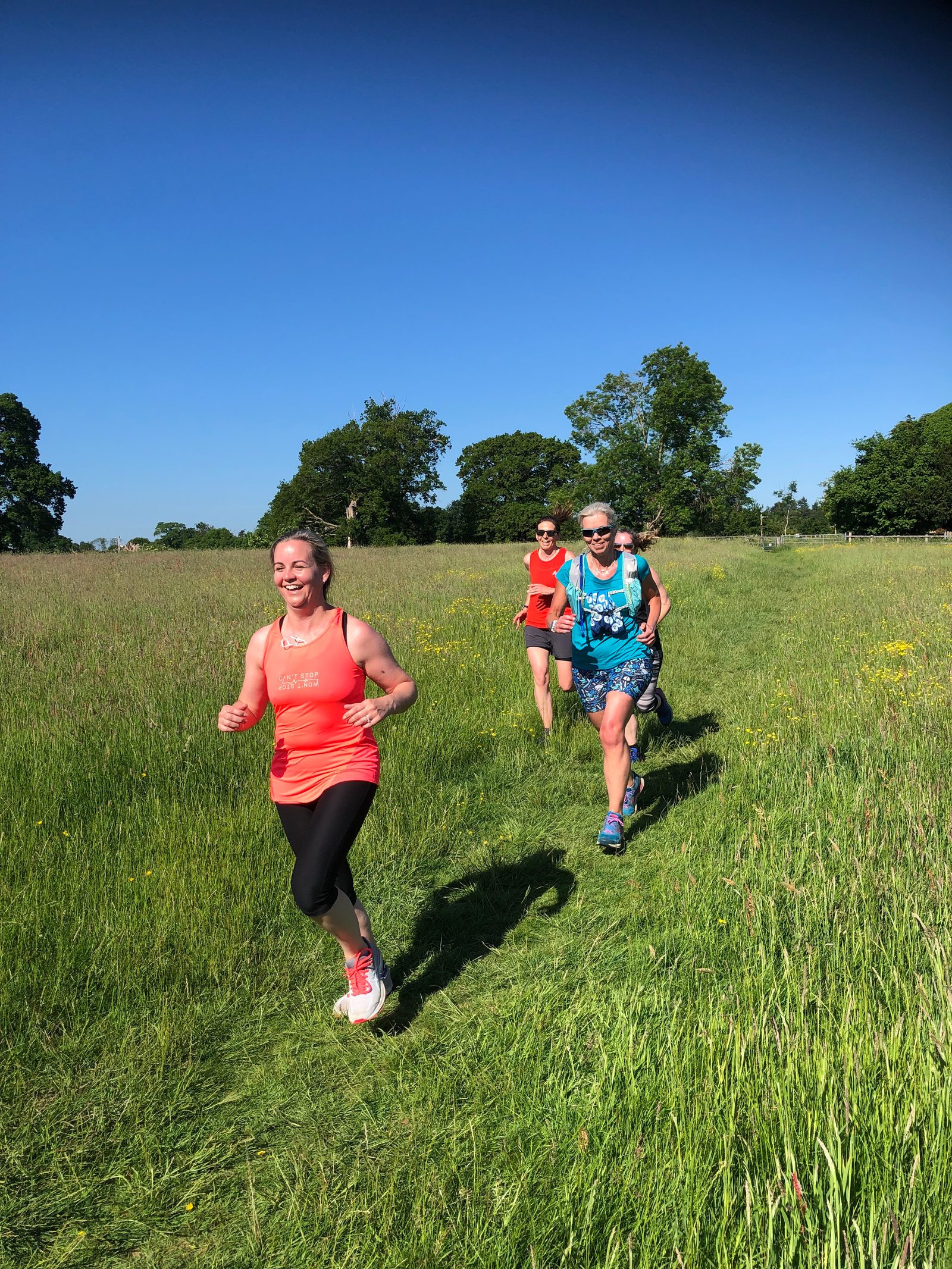 Three people are running on a grassy path through a field under a clear blue sky. The foremost runner is a woman in an orange tank top and black leggings, followed by a woman in a red top and shorts, and a man in a blue tank top and shorts. Trees are in the background.