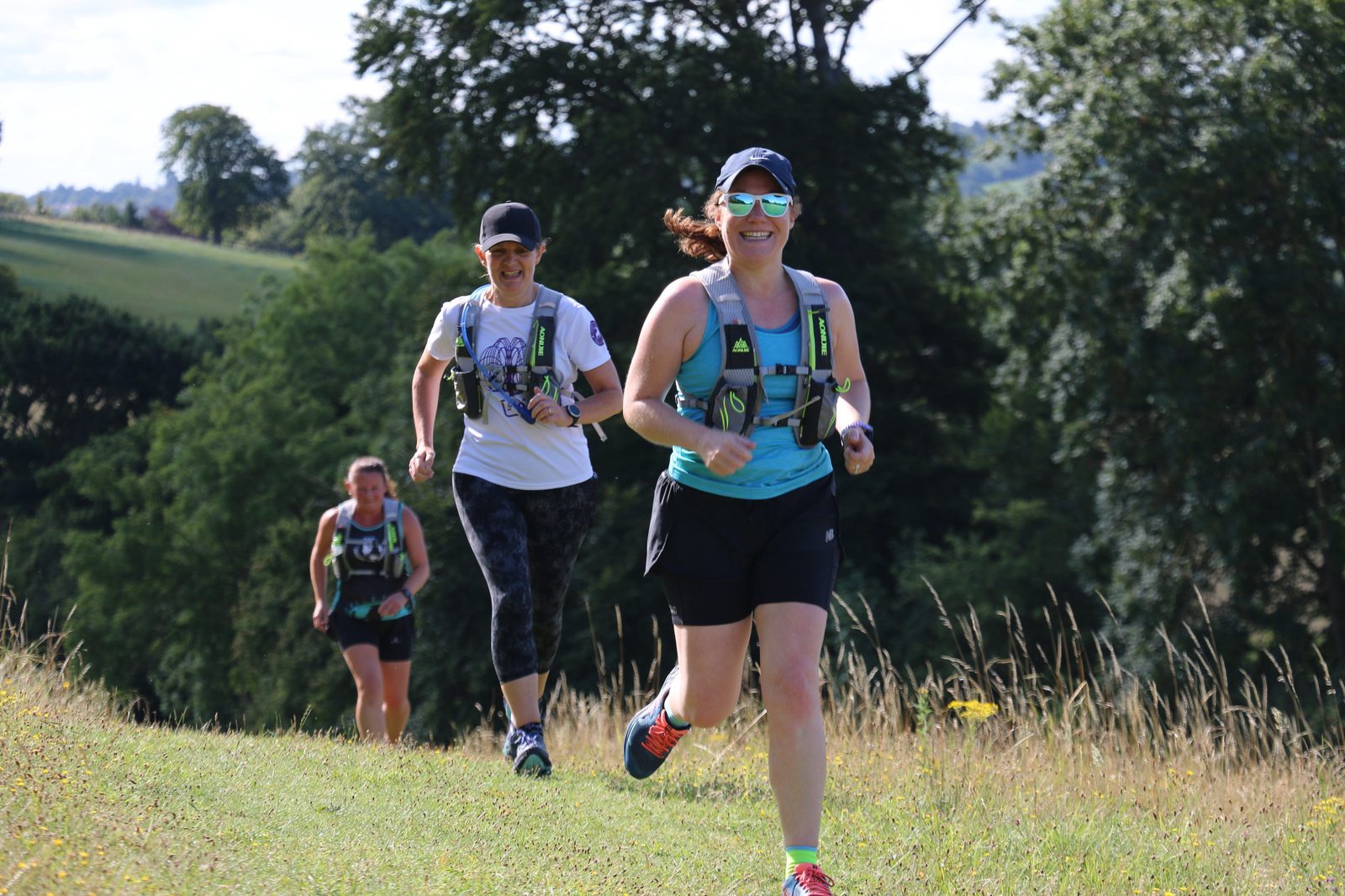 Three people are running on a grassy trail through a scenic landscape with trees in the background. The foreground runner is smiling and wearing a blue tank top, sunglasses, and a hydration pack. The other two runners follow behind, also wearing athletic attire and hydration packs.