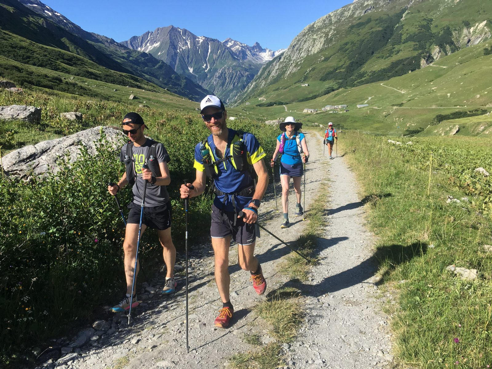 Four hikers trek along a rocky mountain path surrounded by lush green hills and distant snowy peaks under a clear blue sky. All wearing athletic gear and hats, they use trekking poles for support and enjoyment on the scenic trail.