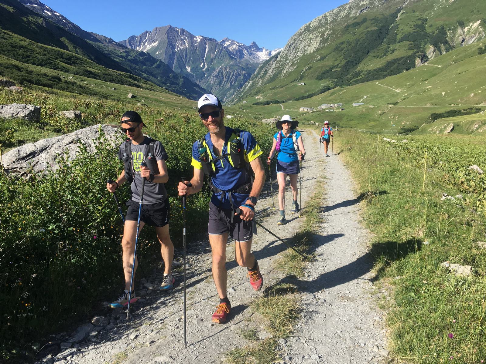 Four hikers with backpacks and trekking poles walk on a dirt trail in a mountainous area with lush green vegetation. They are dressed in athletic wear, and the sky is clear. Snow-capped peaks and rugged cliffs dominate the background.