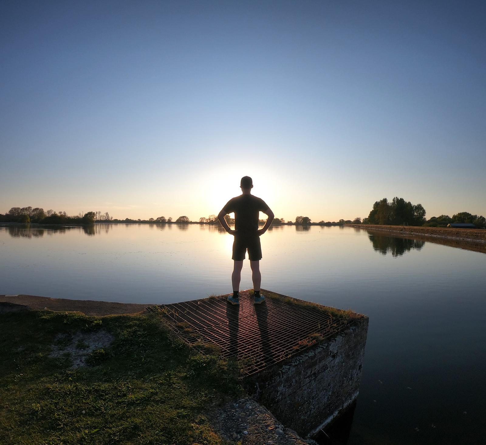 A person stands on a grated platform at the edge of a calm lake, with hands on hips, facing the sunset. The surroundings include grassy areas and trees on the horizon, reflecting on the water's surface. The sky is clear with the sun low, casting a serene glow.