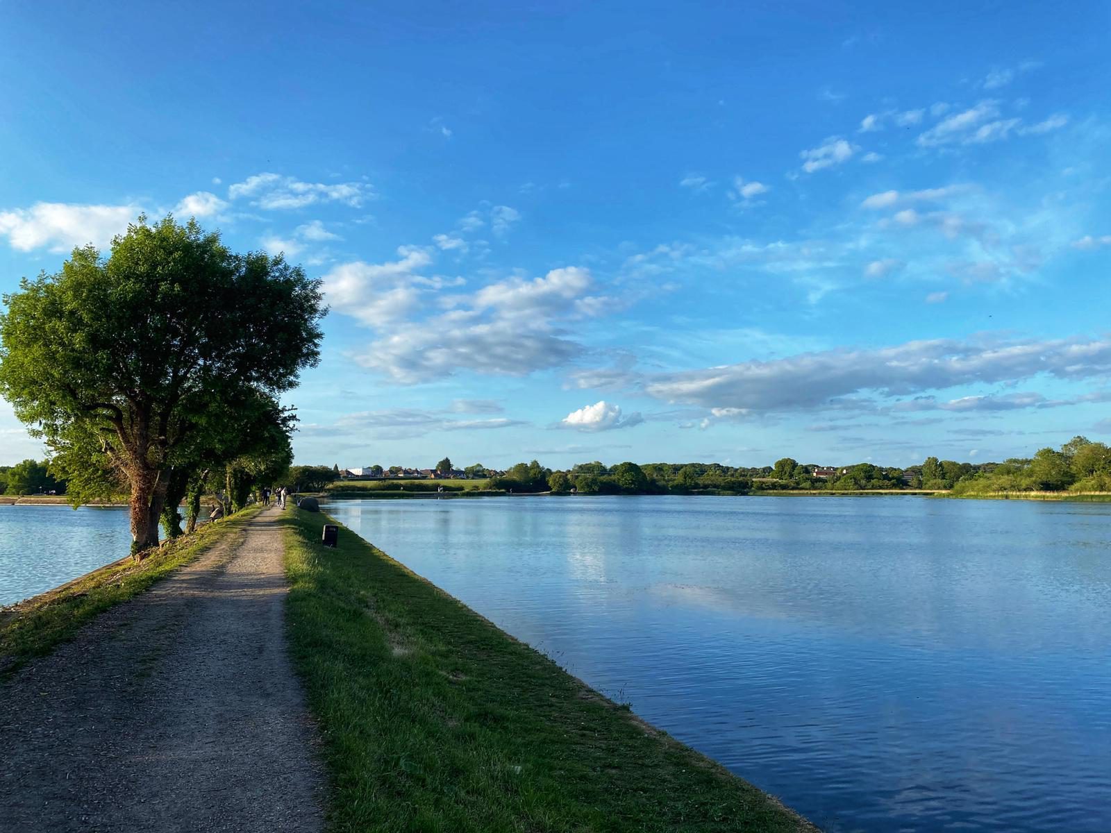 The image shows a tranquil lakeside path bordered by lush green trees on the left and a calm, expansive lake on the right. The sky is bright blue with scattered white clouds, and the sunlight casts a warm glow on the landscape.