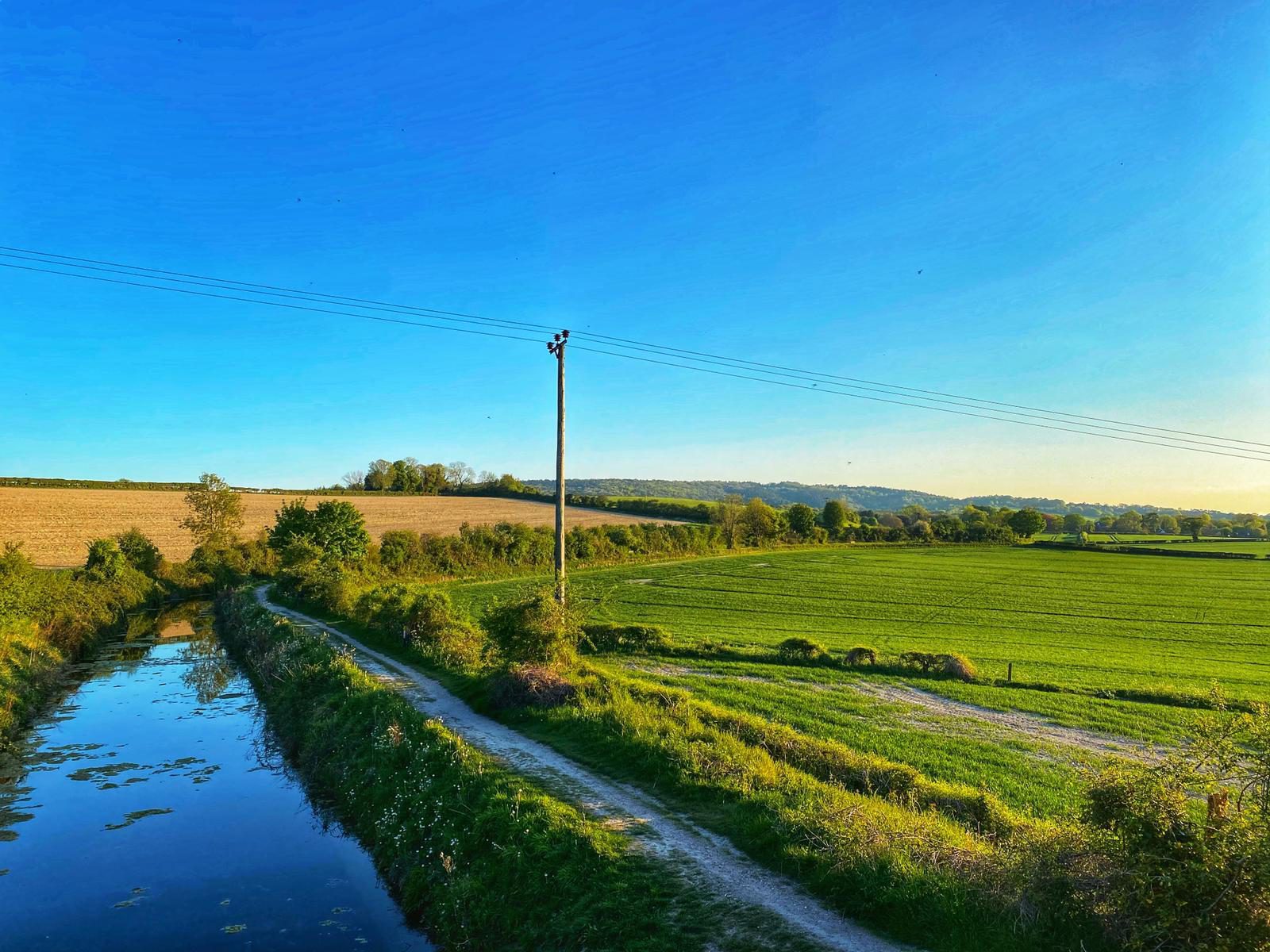 A serene countryside landscape featuring a narrow river running beside a path. On the left, there are fields with a mix of green and brown shades, while the right side showcases lush green fields extending into the distance. A utility pole is seen in the middle. A clear blue sky overhead.